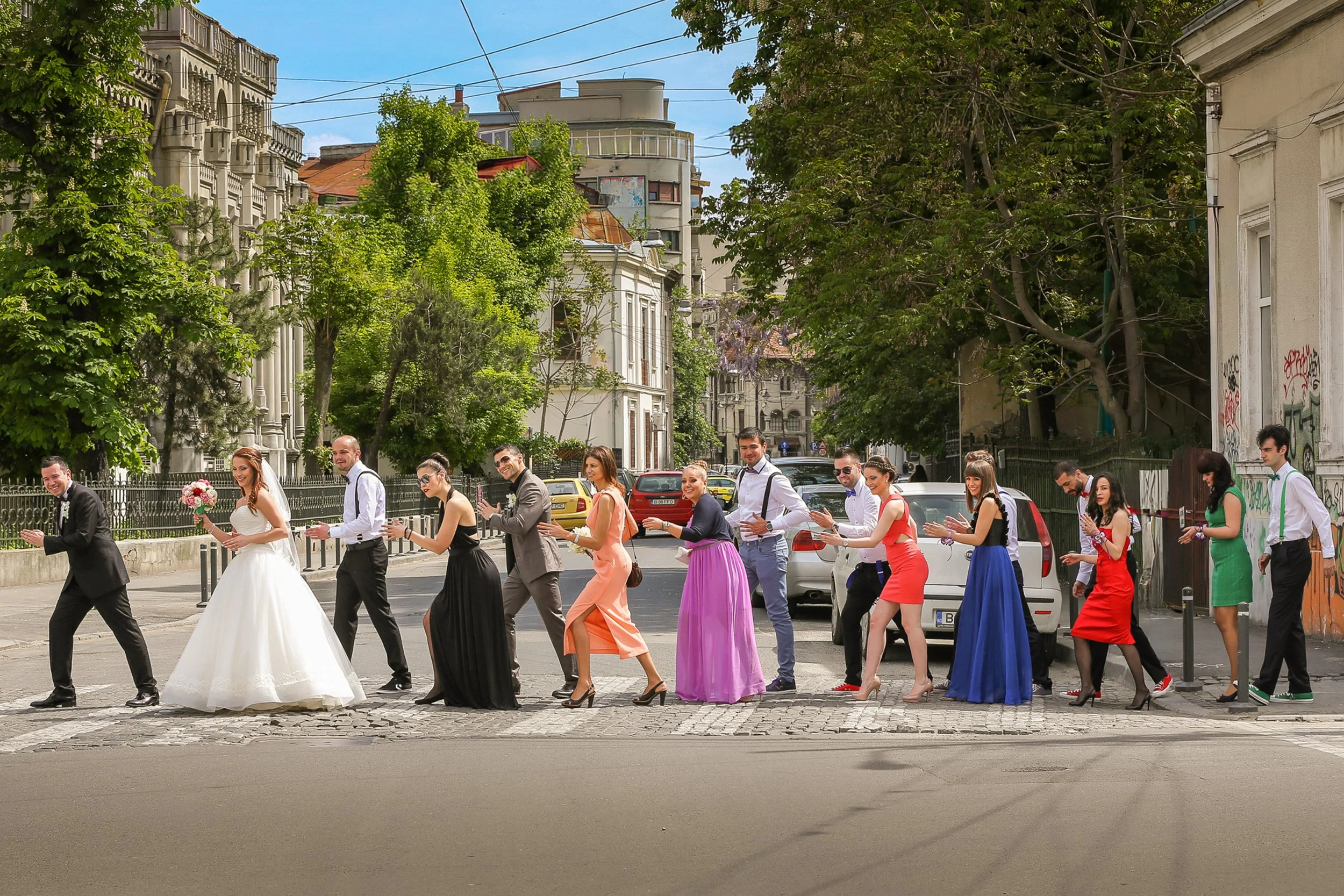A group of people dressed in colorful formal attire, including a bride in a white wedding gown holding a bouquet, walking in a line on a city street with parked cars and buildings, trees, and power lines in the background during daytime.