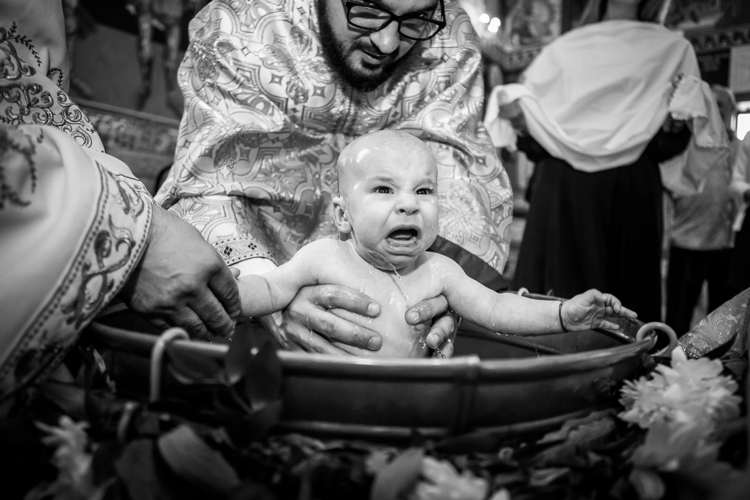 A man with glasses and a patterned shirt giving a baby a bath in a bathtub, and the baby is crying. Other people are visible in the background.