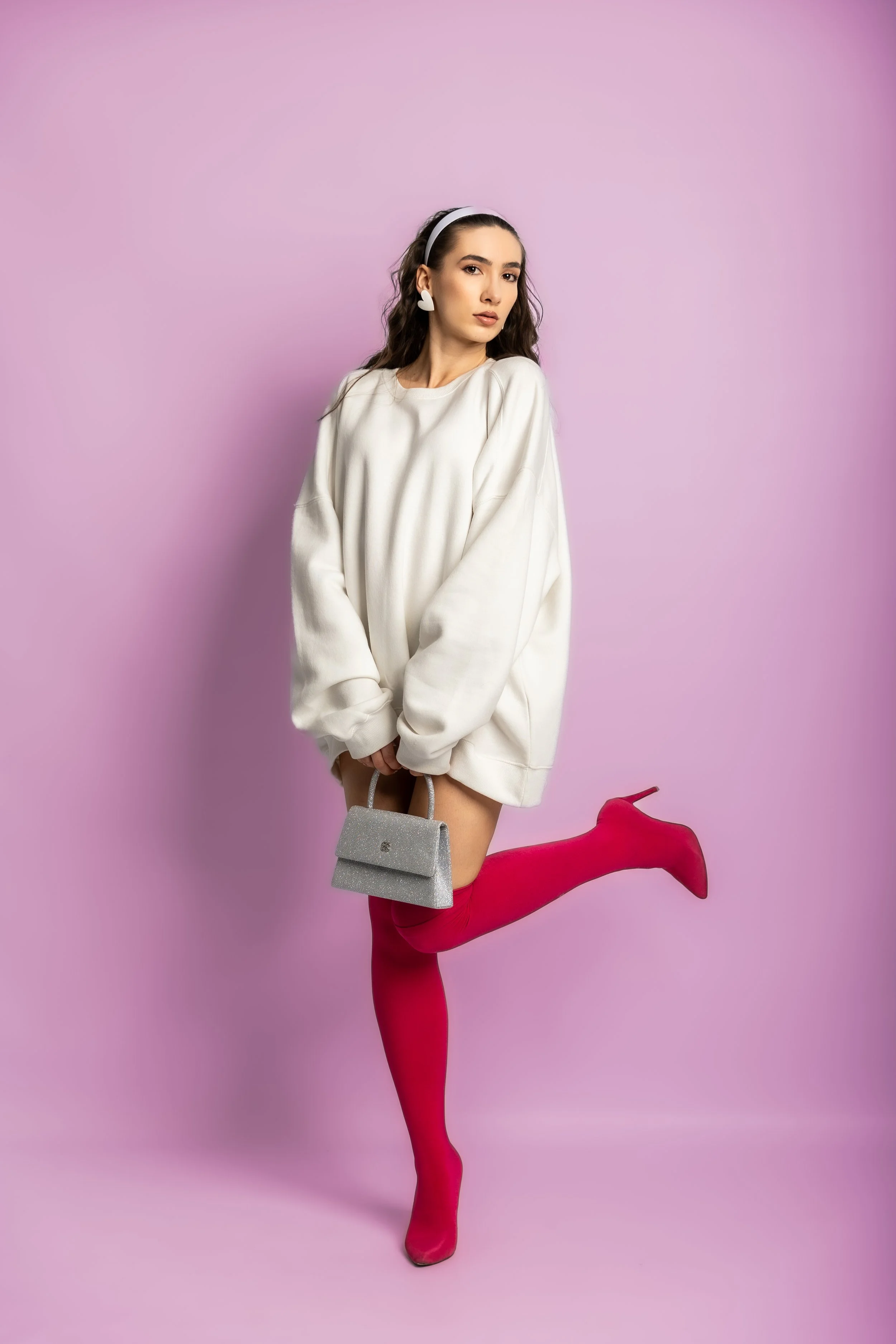 A young woman with long curly hair wearing a white sweatshirt, pink thigh-high stockings, and holding a silver handbag, is posing with one leg lifted, against a pink background.