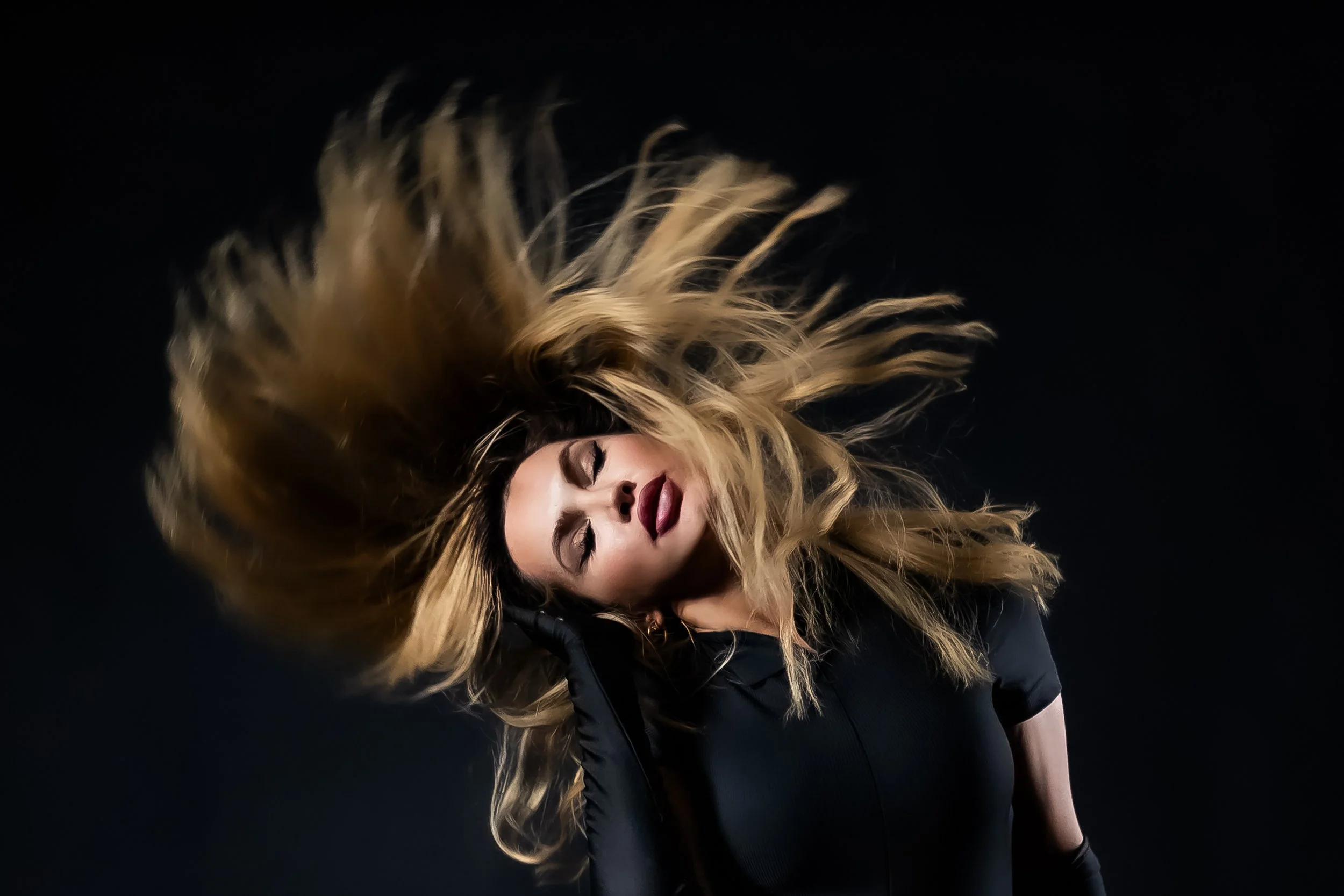 A woman with long blonde hair in motion, wearing a black jacket, against a black background.