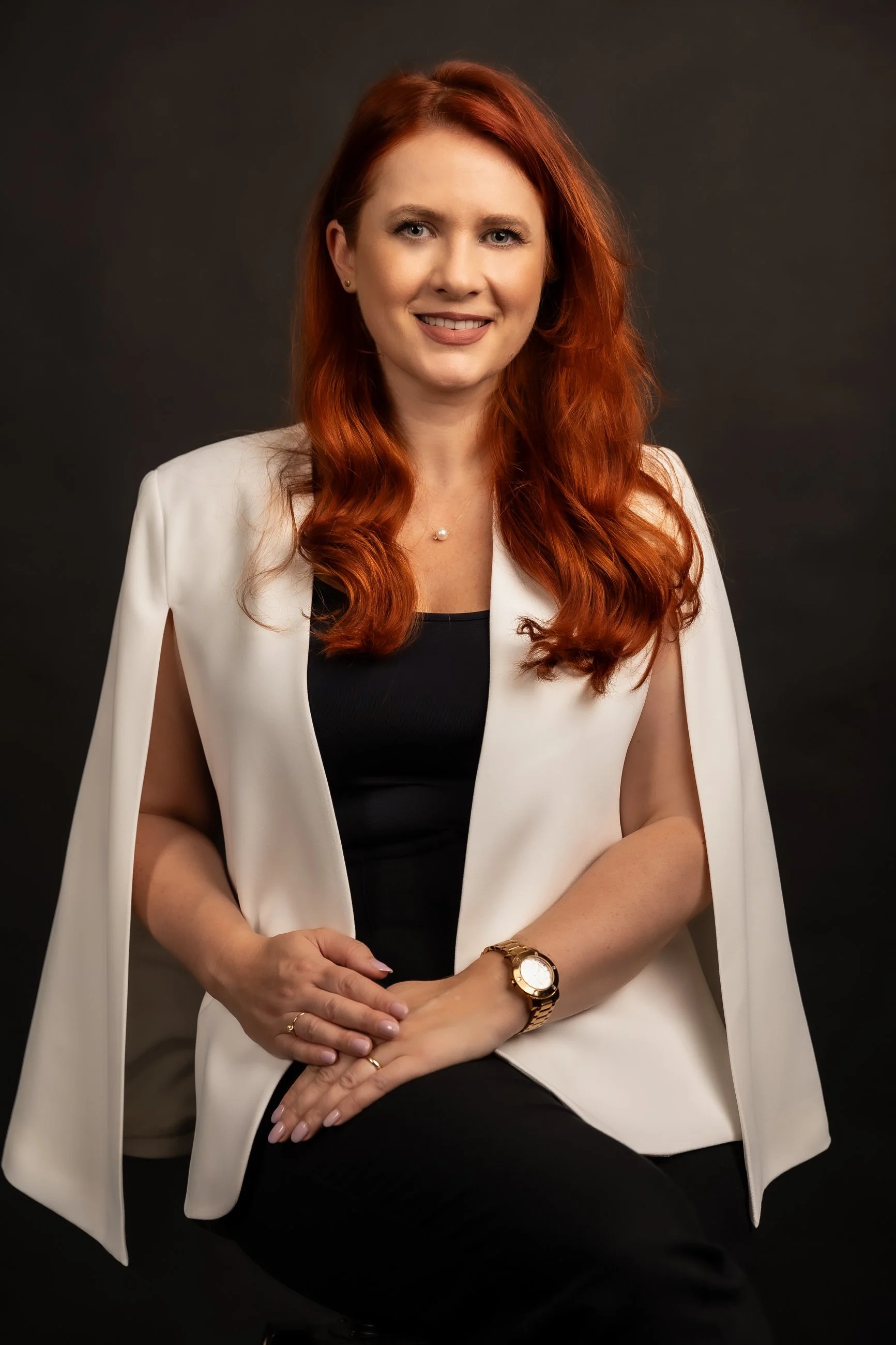 Portrait of a woman with red hair, wearing a black top, white blazer, and a gold wristwatch, sitting with hands crossed, against a dark background.