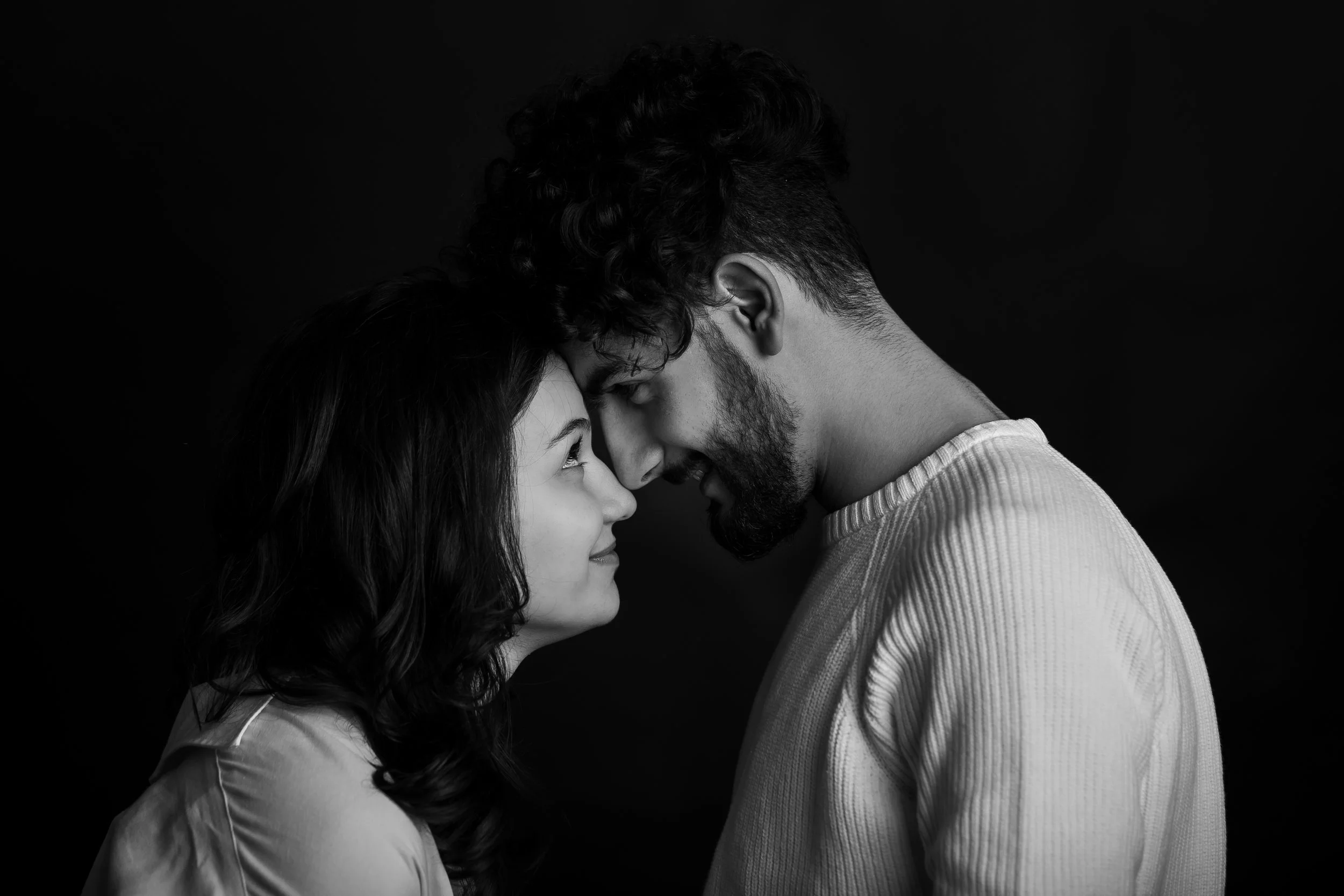 A black and white photo of a man and woman with their foreheads touching, smiling and looking into each other's eyes, with a dark background.