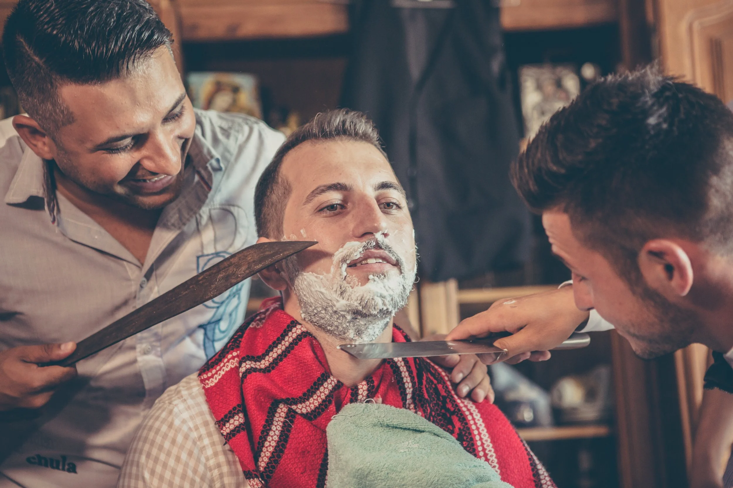 Three men, two with dark hair and one with gray hair, are gathered around a barber's chair with the man in the center having shaving foam on his face and a red and black striped towel around his shoulders, as they prepare for or are in the process of