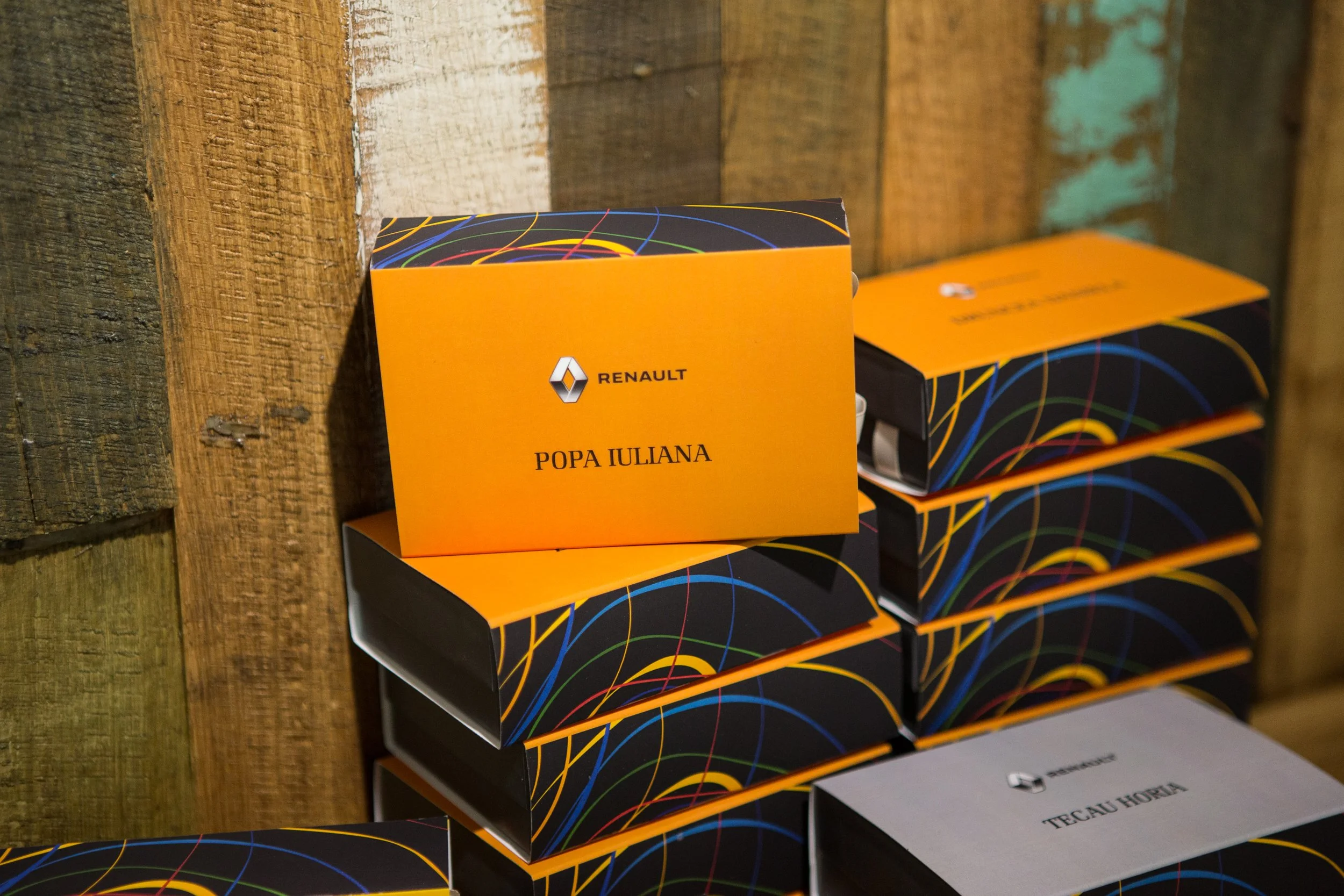 Close-up of a stack of black and orange packages with colorful swirl patterns, on a wooden surface. One package has a label reading 'POP IULIANA' and features the Renault logo.