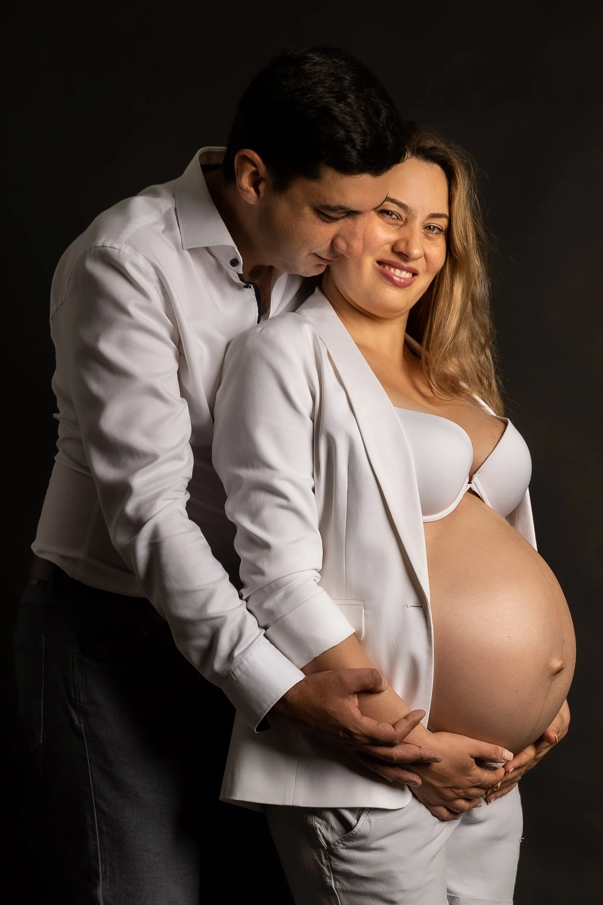 A pregnant woman wearing white lingerie and an open white blazer, smiling, with a man behind her leaning in close and touching her belly, both dressed in white shirts, against a black background.