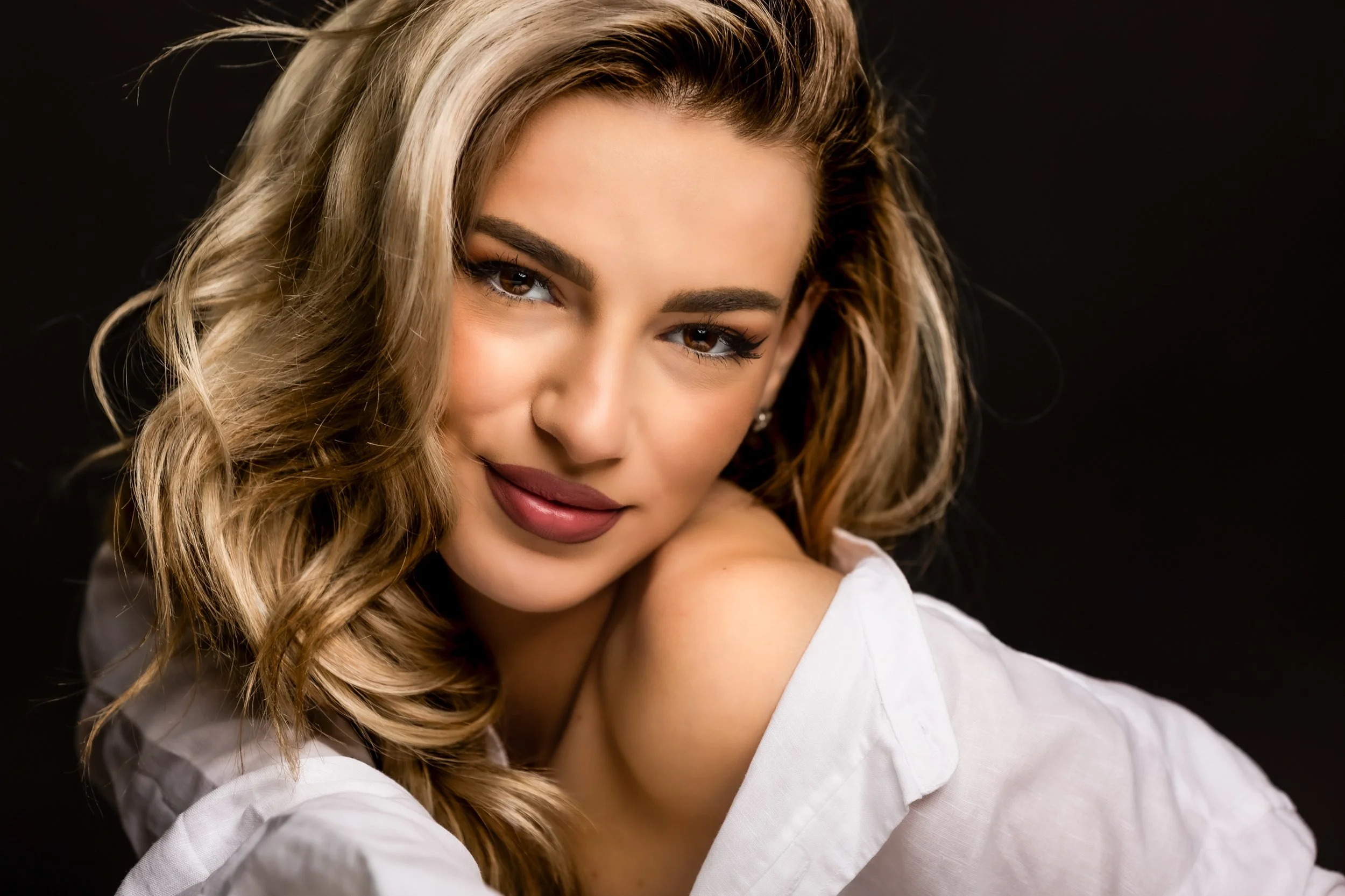 A woman with wavy blonde hair, wearing makeup, smiling softly with her face slightly turned, against a black background.