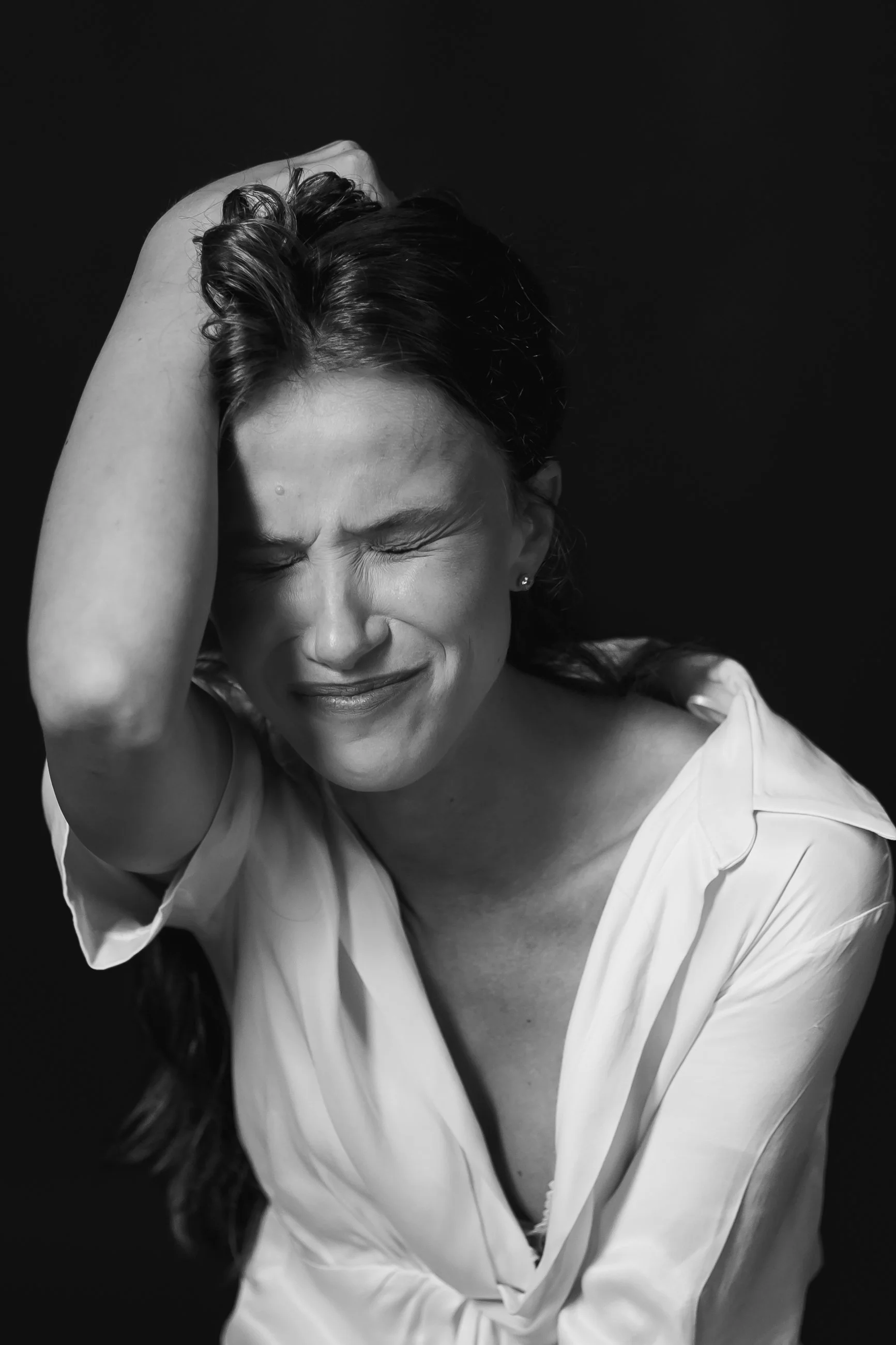 Black-and-white photo of a woman with closed eyes, smiling, holding her head with one hand, wearing a light-colored shirt, against a dark background.