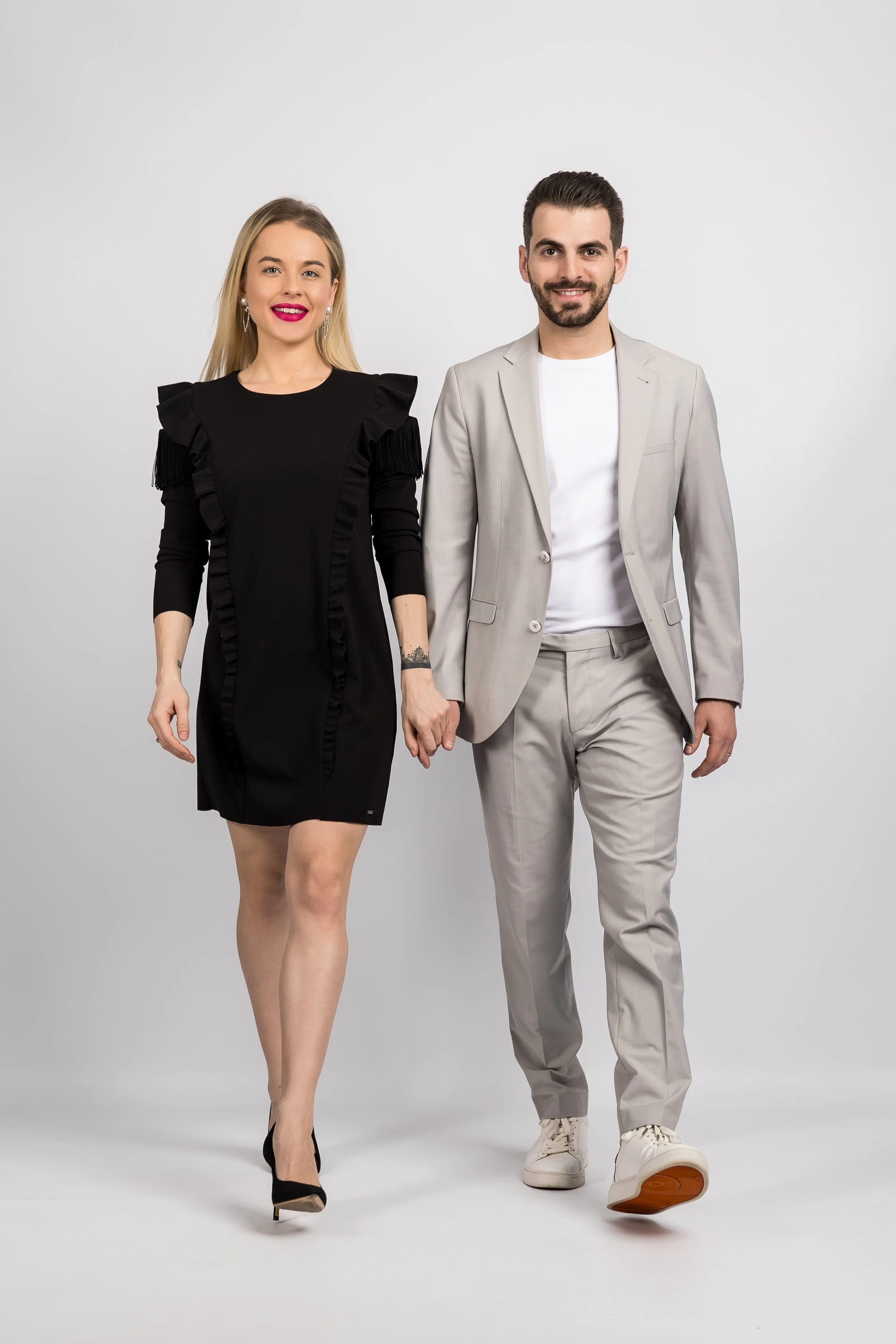 A man and woman walking hand in hand, both dressed in stylish outfits, with the woman wearing a black dress with ruffle details and the man in a light suit with white sneakers, on a plain white background.