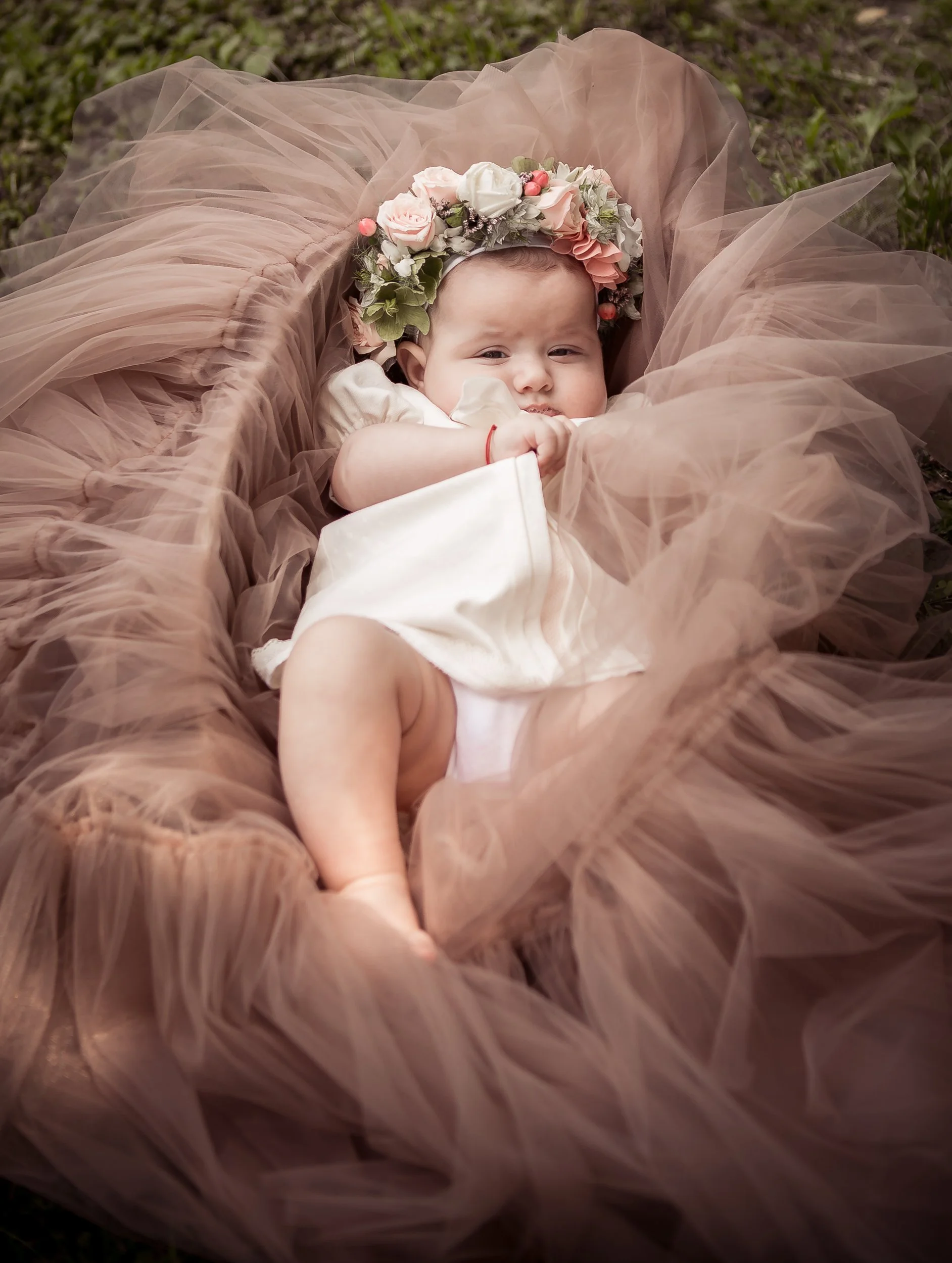 A baby lying on a soft pink tulle blanket, wearing a floral crown, outdoors on grass.