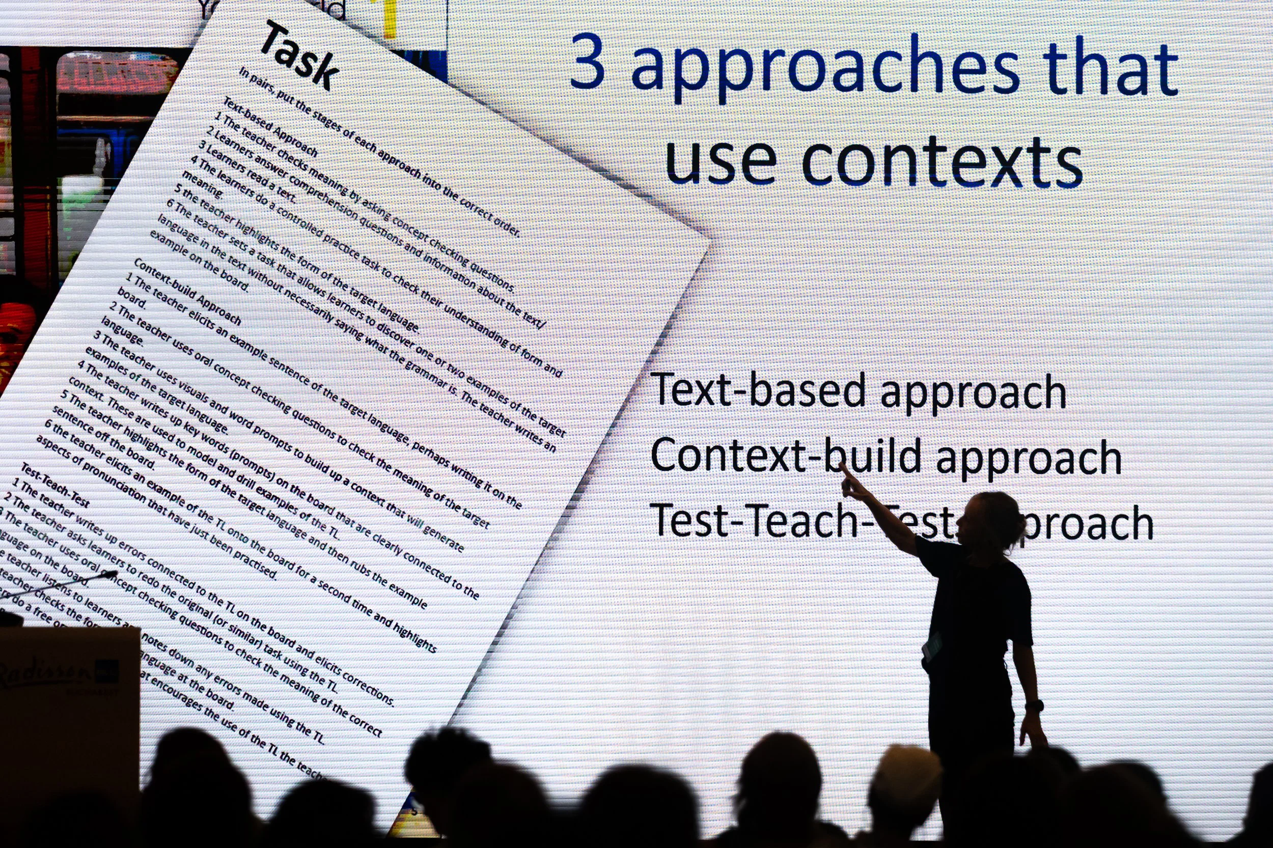 A large presentation slide with text about different approaches to understanding and teaching language, including task and context-building approaches. A speaker is pointing at the slide with a silhouette visible against the screen, while an audience