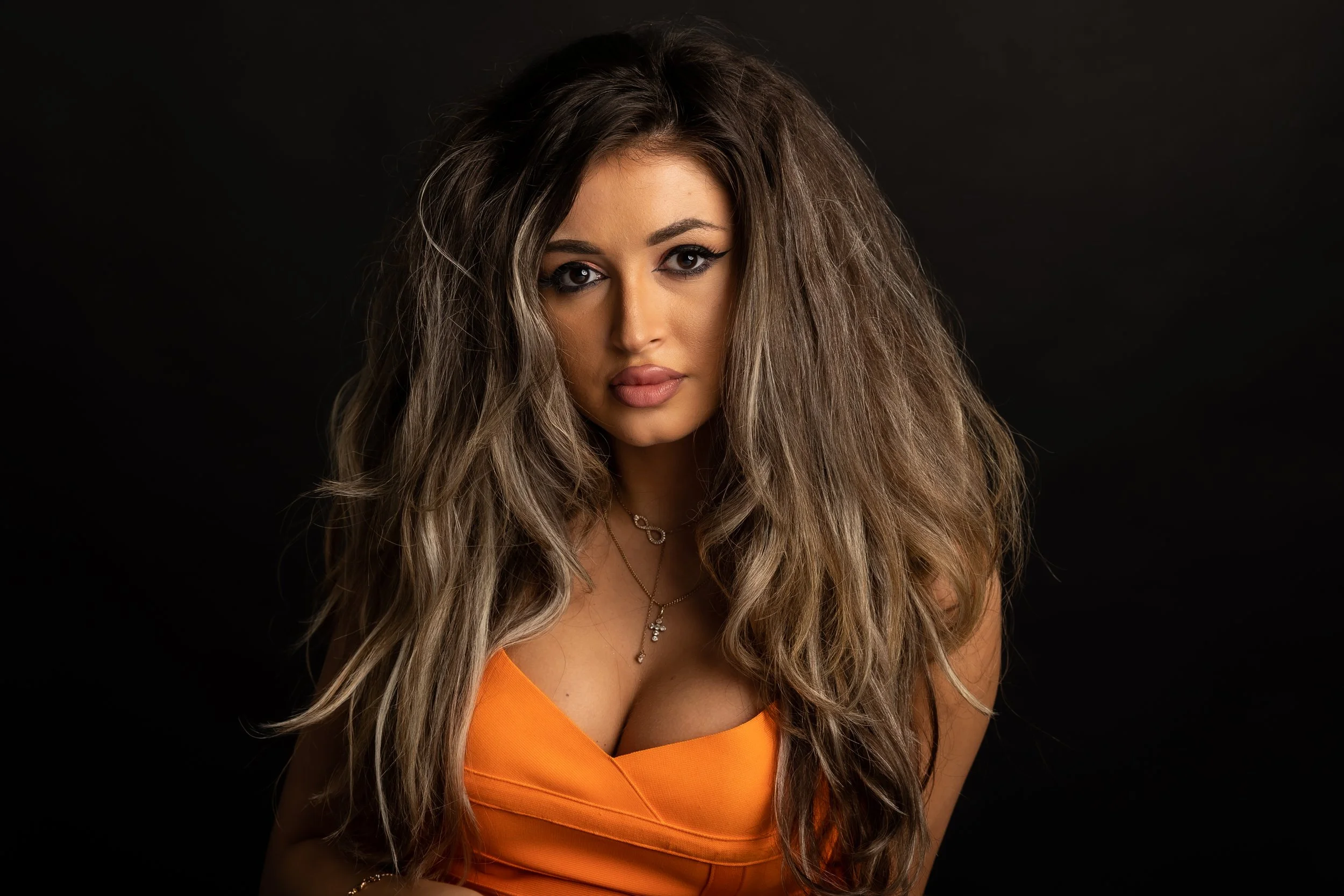 A young woman with long, wavy hair and dark eye makeup wearing an orange top and a necklace with a cross pendant, posed against a black background.
