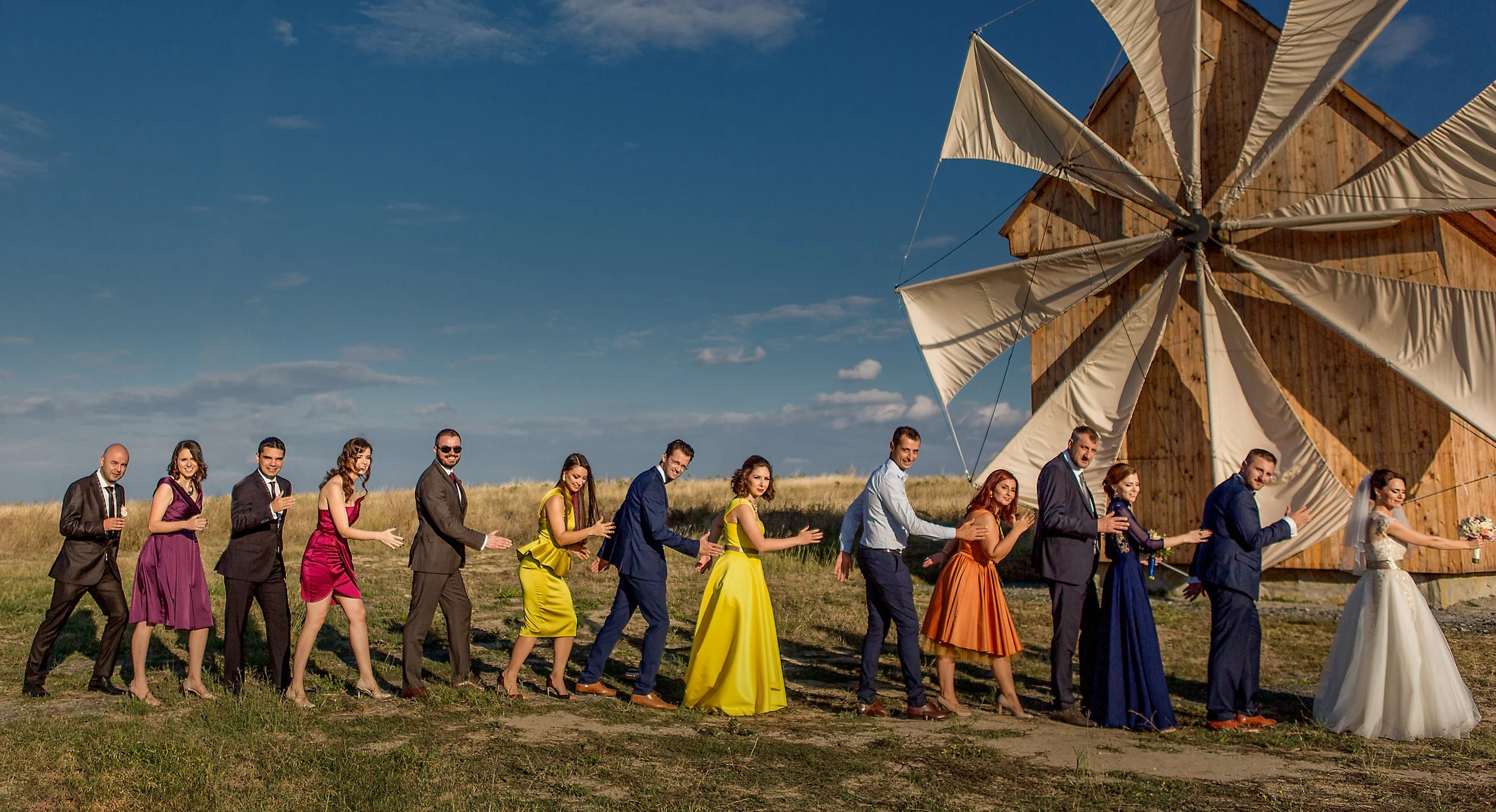 A line of wedding guests in colorful dresses and suits walking outdoors on a grassy field, following a bride holding a bouquet, near a wooden windmill structure with sails, under a partly cloudy blue sky.
