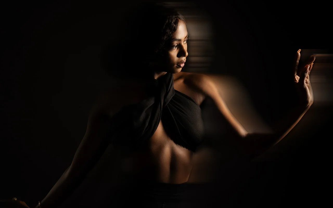 A woman with medium skin tone and short hair wearing a black top, looking to the side with her hand gently raised, dramatic low-light photography.