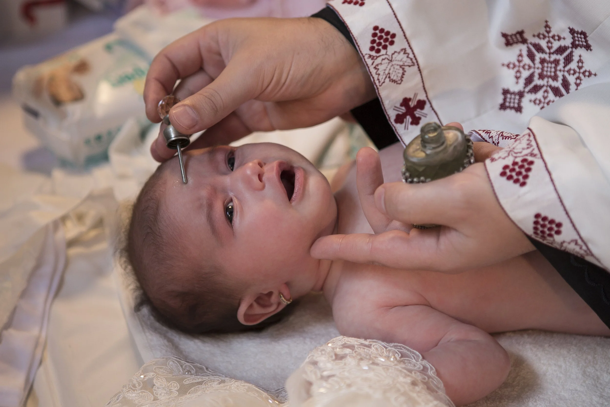 A baby lying down on a bed receiving a medical procedure, with a healthcare professional using a syringe on the baby's forehead, in a setting decorated with holiday or winter-themed embroidery clothing.