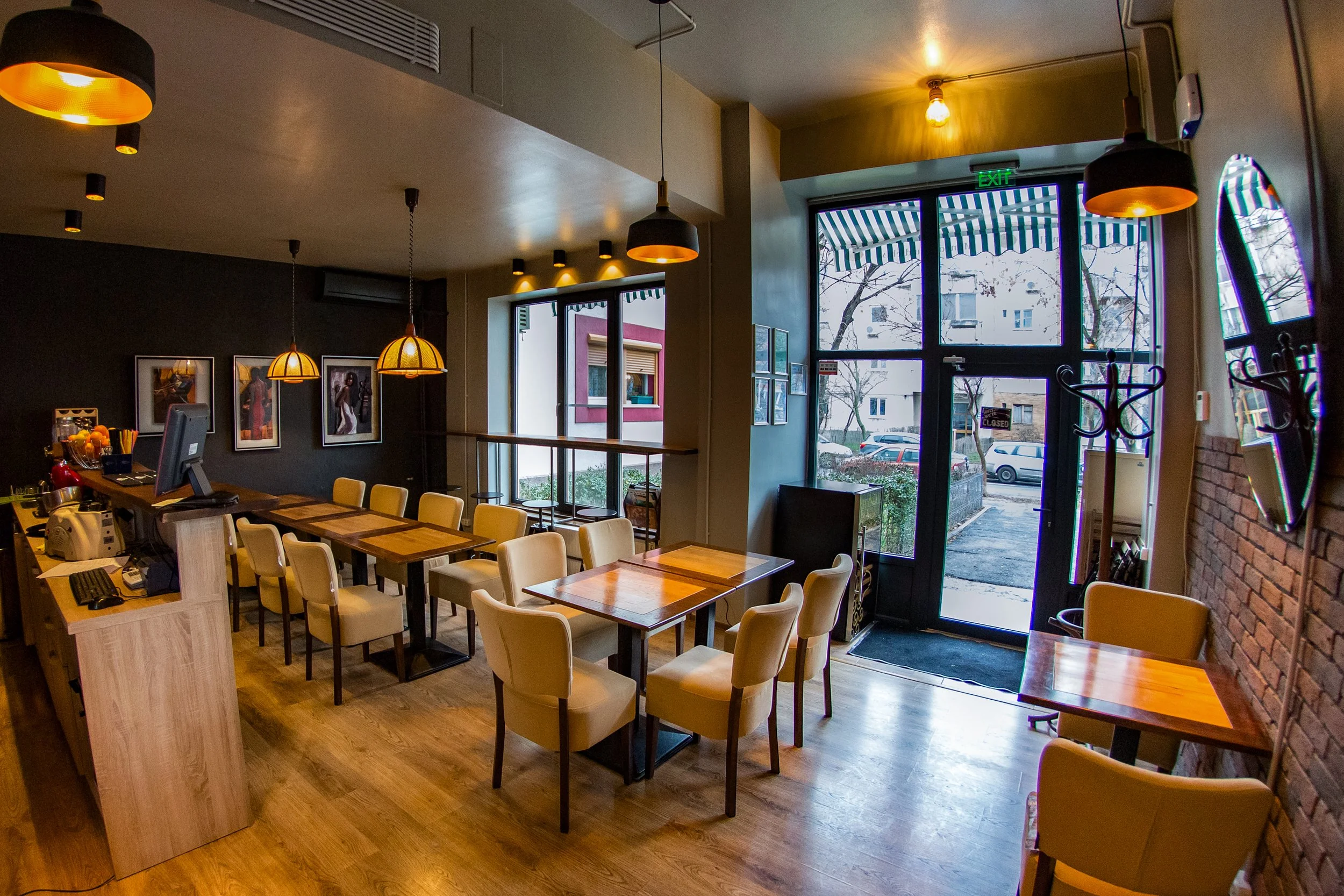 Interior of a cozy cafe with wooden tables and beige chairs. There are hanging pendant lights with warm yellow illumination, framed pictures on the dark accent wall, large windows with striped awnings showing snow-covered outdoors, and a metal coat r