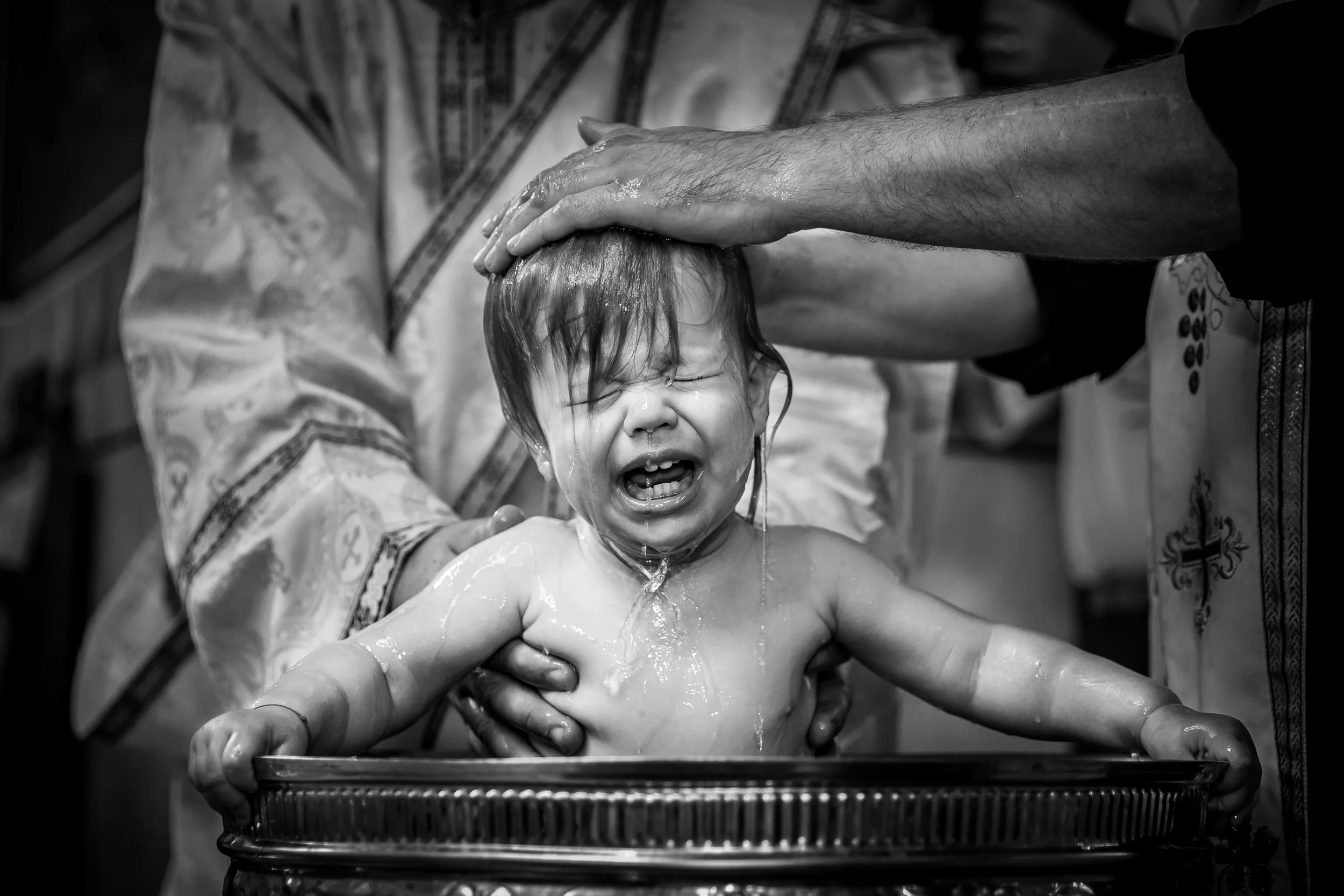A young child is being baptized in a religious ceremony, with a person pouring water over the child's head. The child is crying and adult's hand supports the child's head during the baptism.