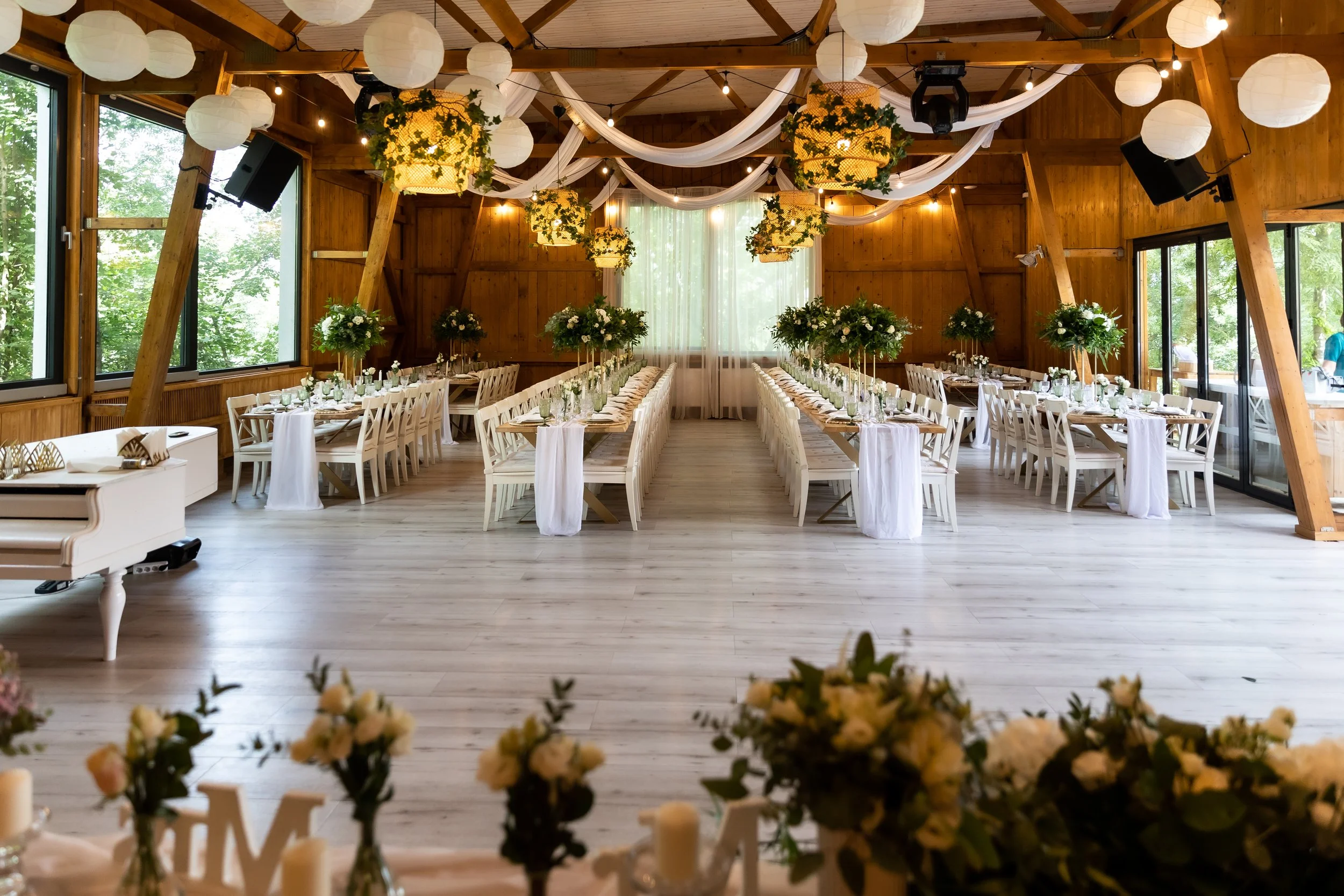 A decorated indoor event space with long rectangular tables arranged in a U-shape, adorned with floral centerpieces, white tablecloths, and glassware. The room features wooden walls and ceiling, large windows showing greenery outside, and white paper