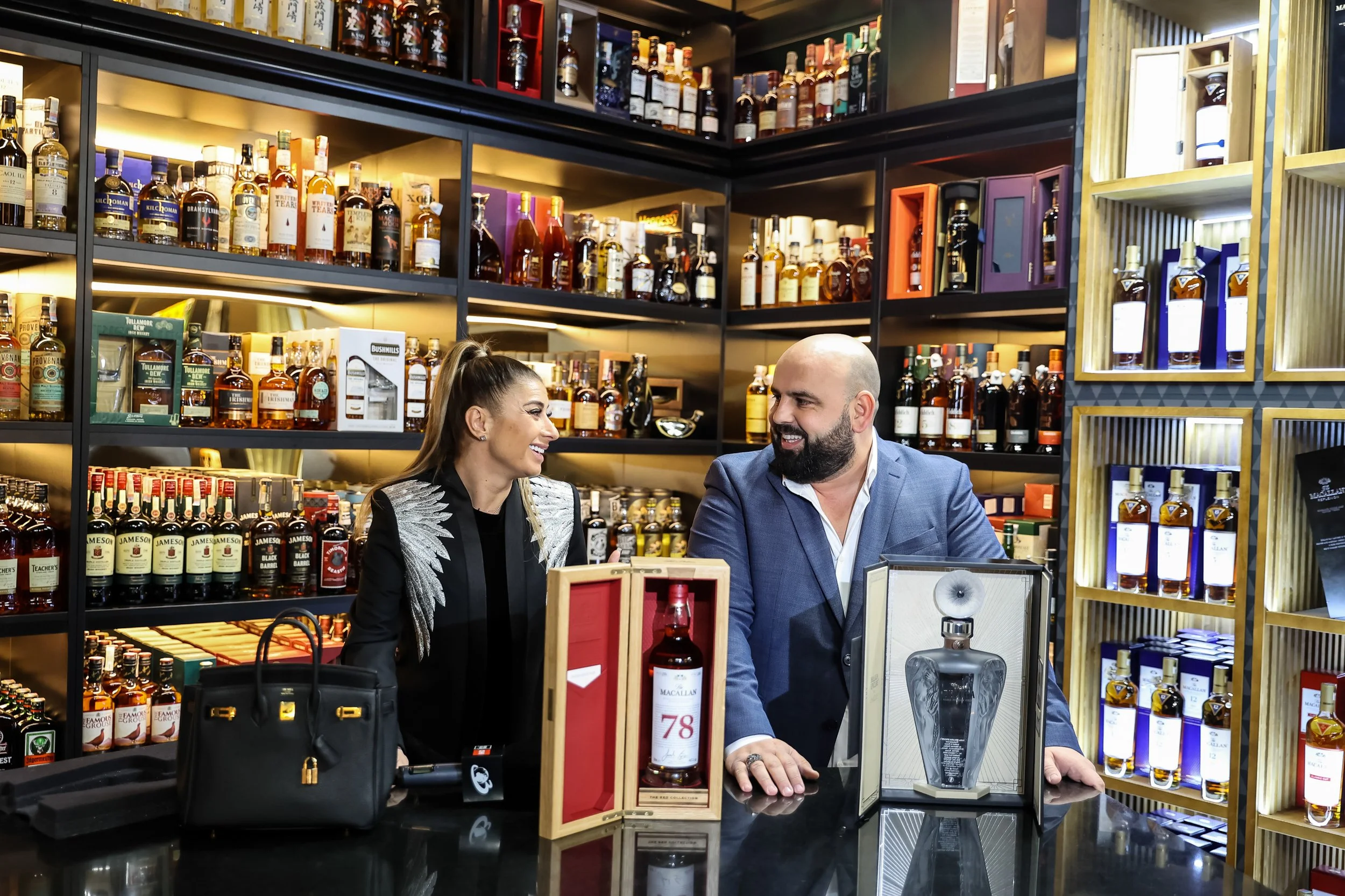 A man and a woman smiling at a counter in a liquor store with shelves of whiskey bottles behind them.