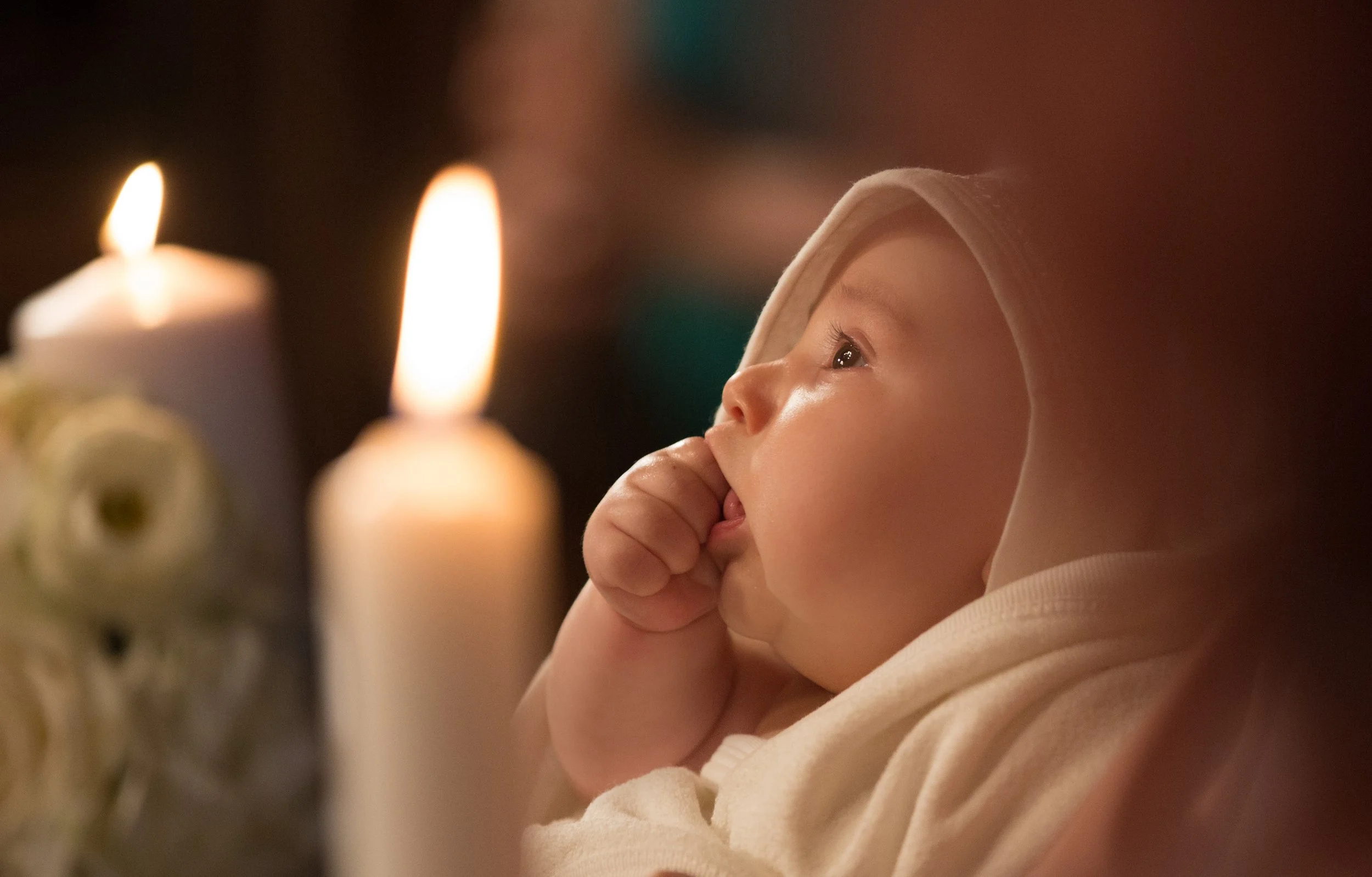 A young child with a hoodie looks at lit candles with a thoughtful expression in a dark room.