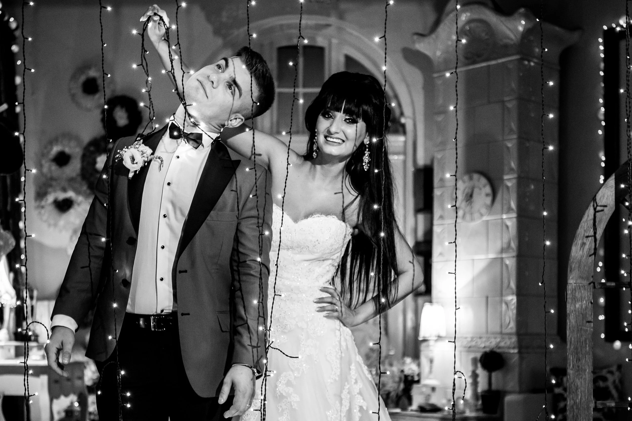 A black and white photo of a newlywed couple standing indoors during their wedding reception, behind decorative string lights. The groom wears a tuxedo with a bow tie, and the bride wears a strapless wedding gown with earrings. They are smiling and l