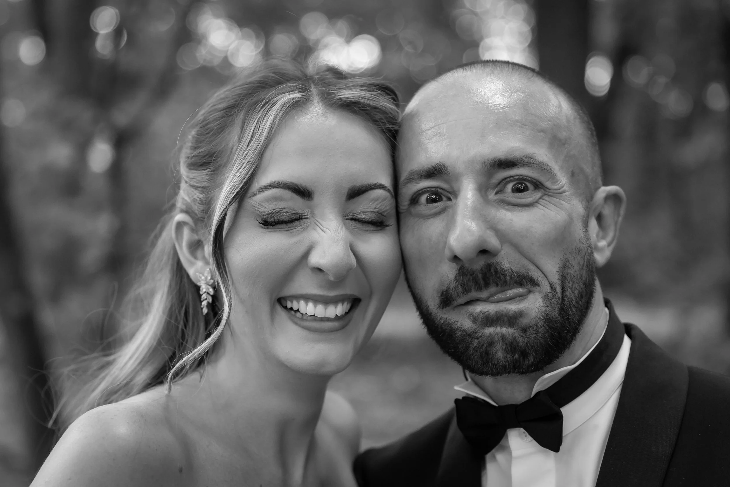 Black and white close-up photo of a smiling woman with long hair and a man with a bald head, both dressed in formal attire, with a blurred outdoor background.