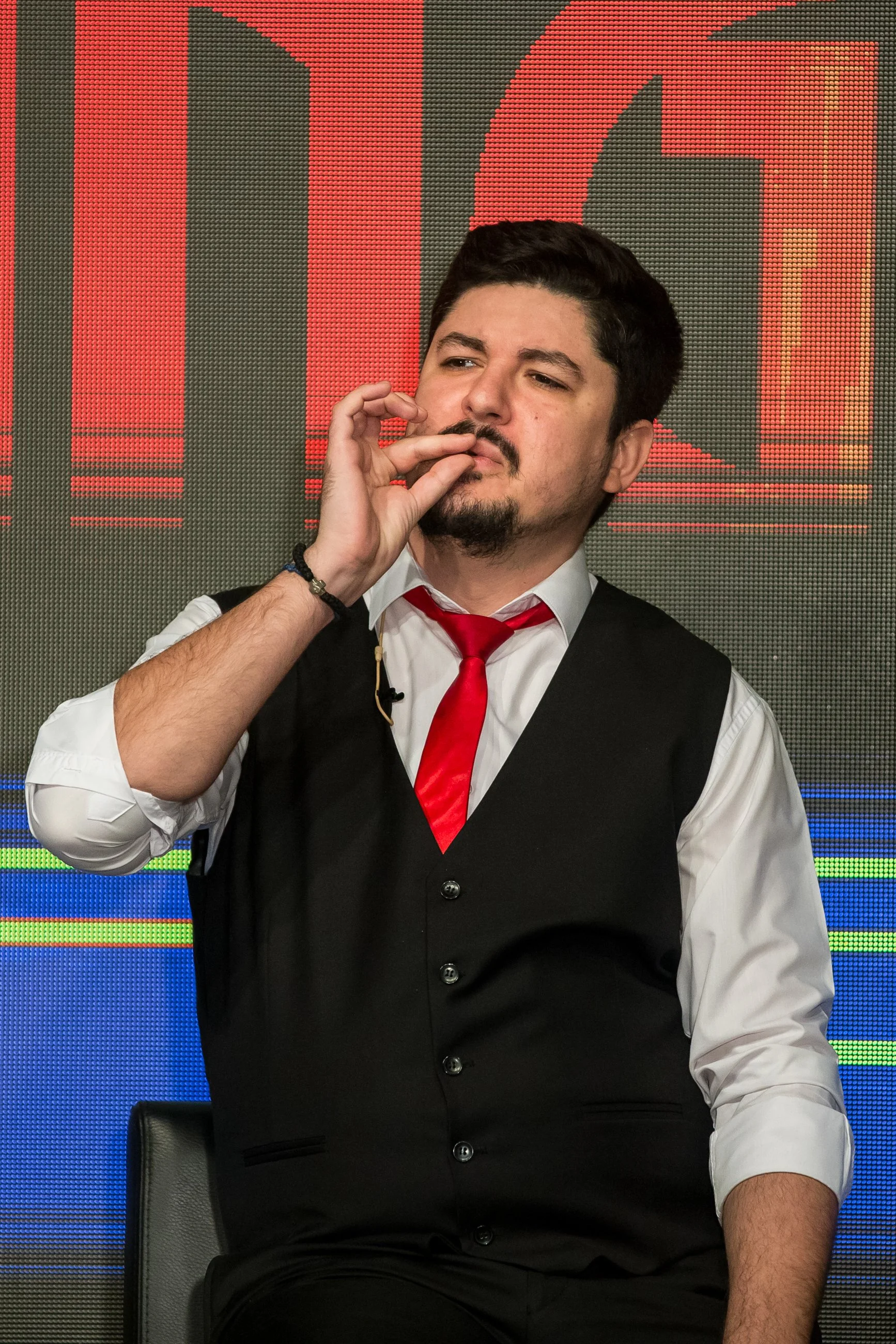 A man wearing a formal vest, white shirt, and red tie sitting in front of a digital screen with red and blue graphics, appearing deep in thought.