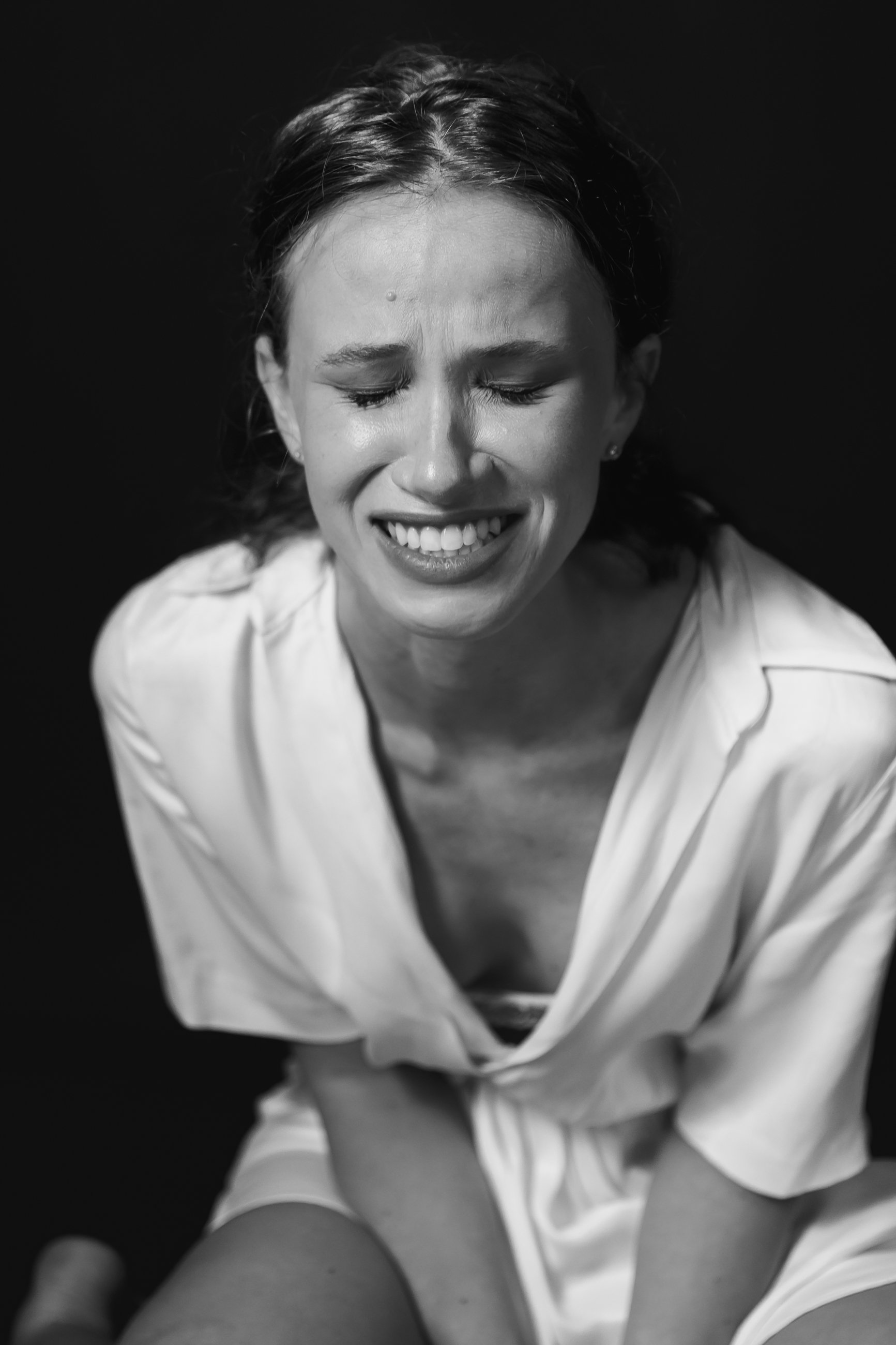 A woman with wet hair, eyes closed, smiling, wearing a light shirt and earrings, against a black background.