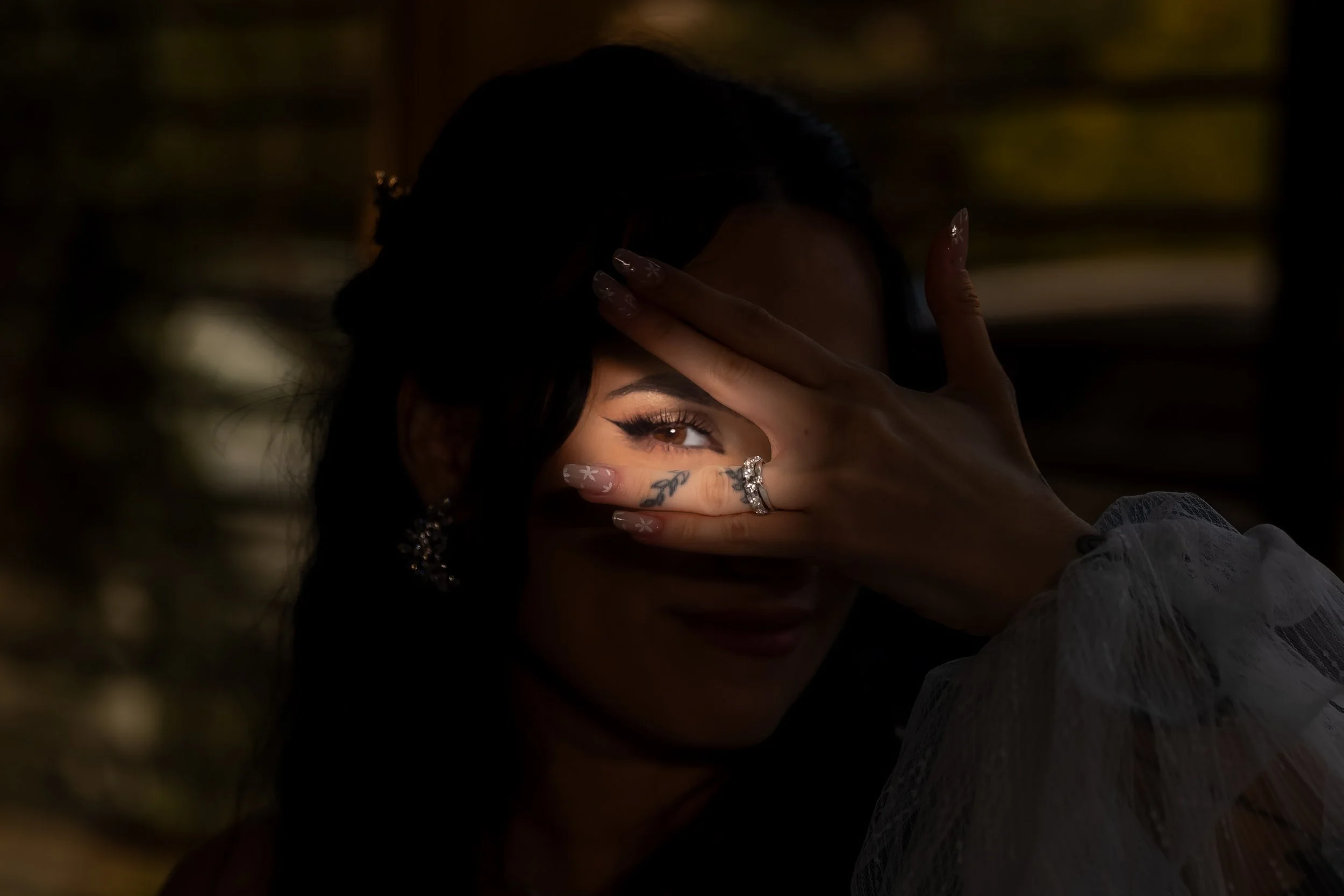 A woman with dark hair and makeup partially covers her face with her hand, showing rings and decorated nails, in a dark, moody setting.