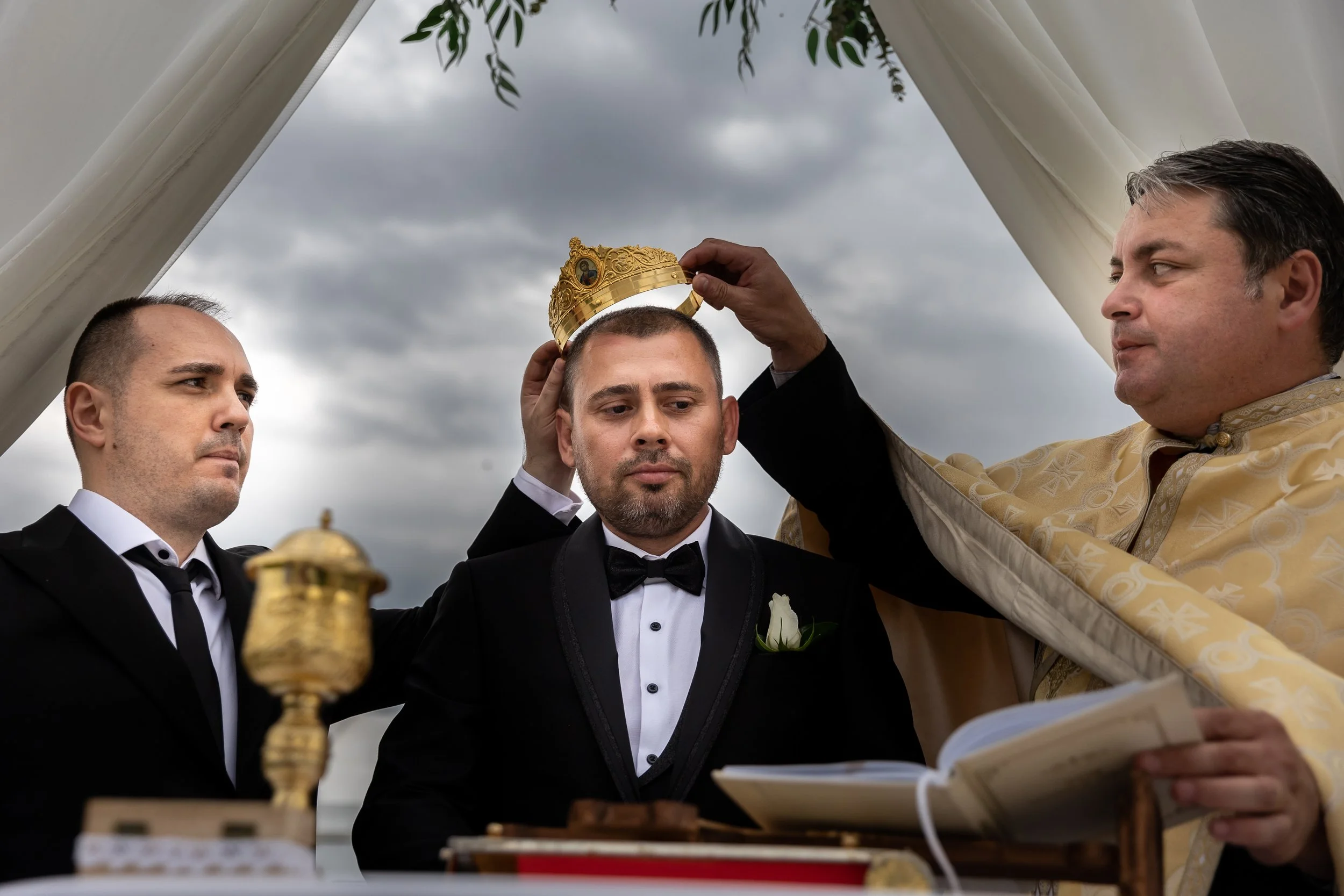 A man is being crowned with a gold crown during an outdoor ceremony under cloudy skies, with two other men in formal attire assisting him.