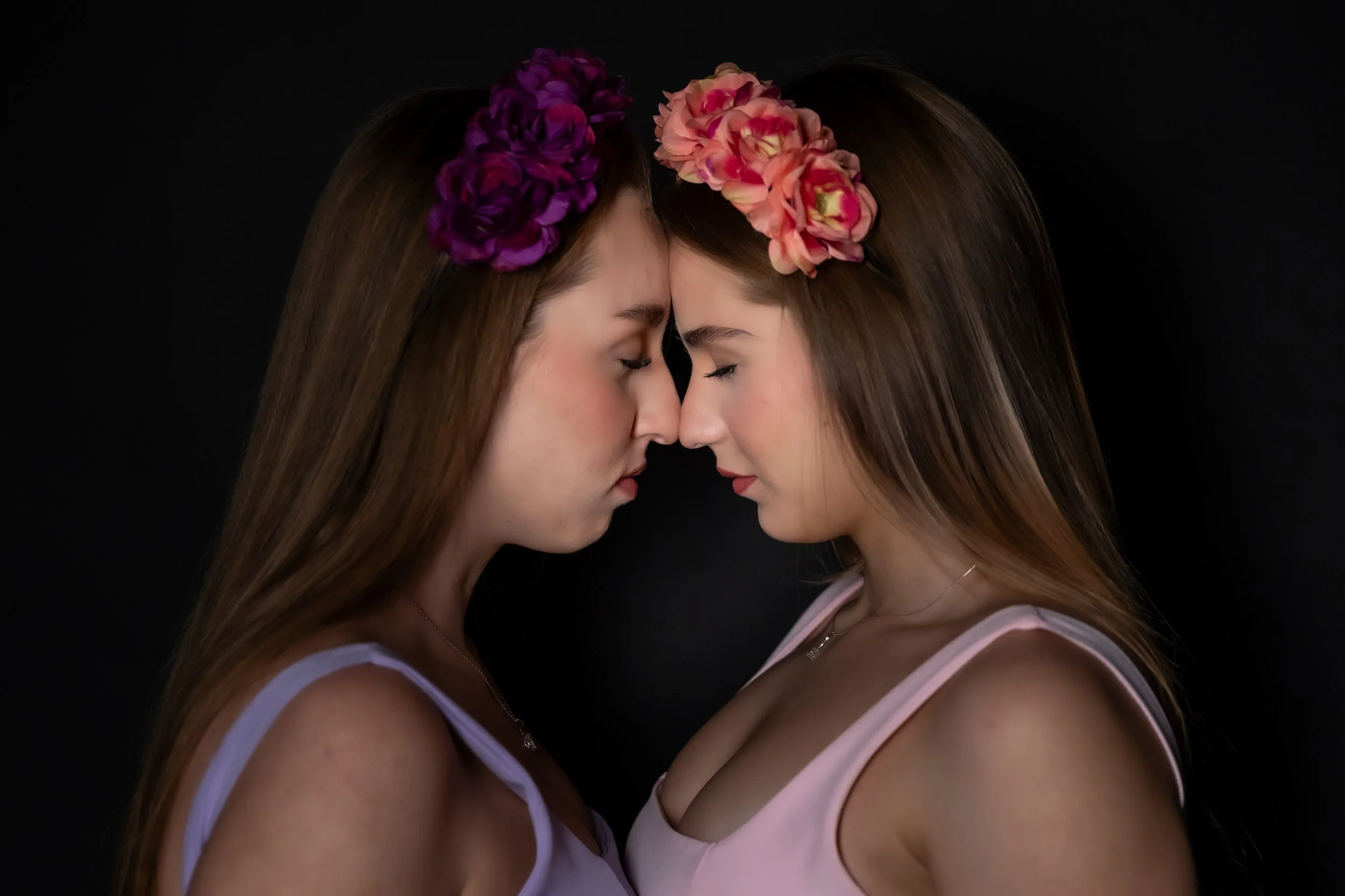Two women with long hair and floral headbands touching foreheads against a black background.