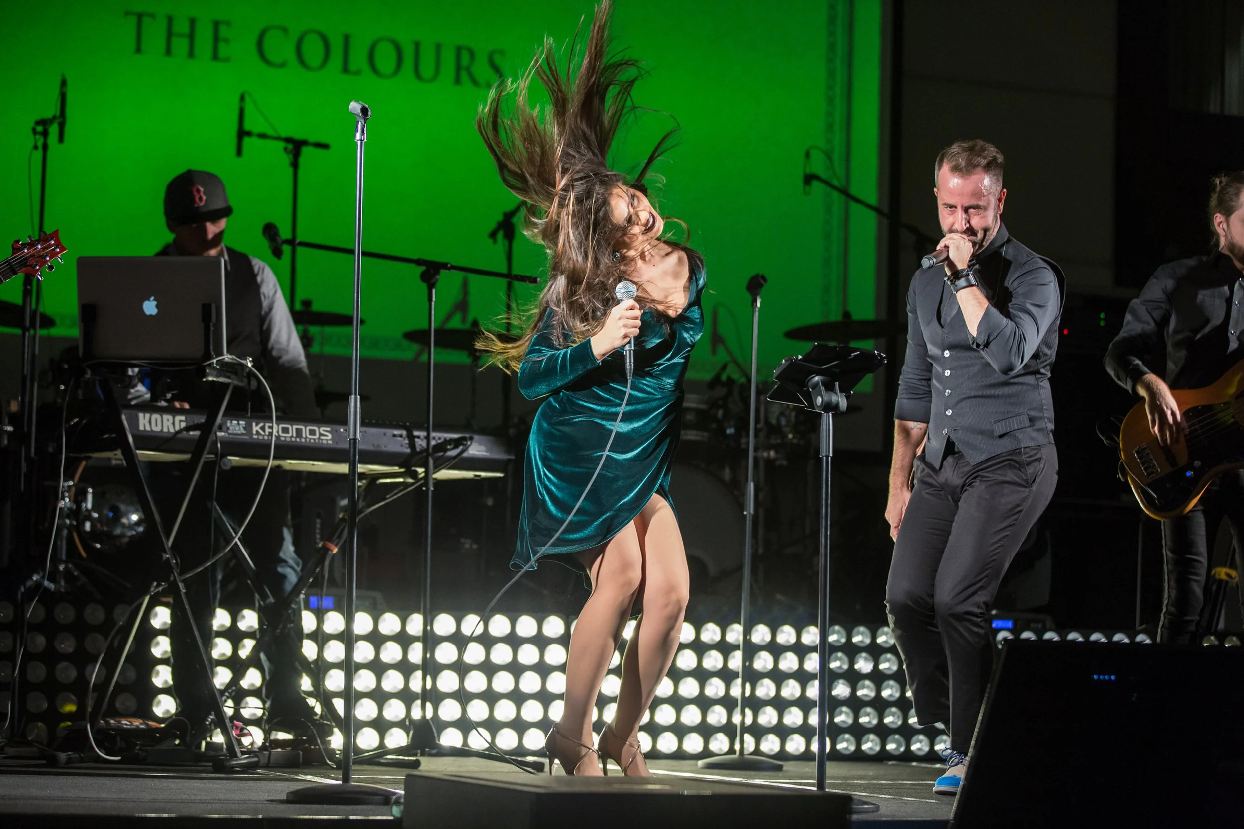 Musicians and singers performing on stage with a green backdrop that reads "The Colours." A woman in a silky teal dress is dancing and singing into a microphone, with her hair flying in the air. A man in a dark shirt and jeans is holding a microphone