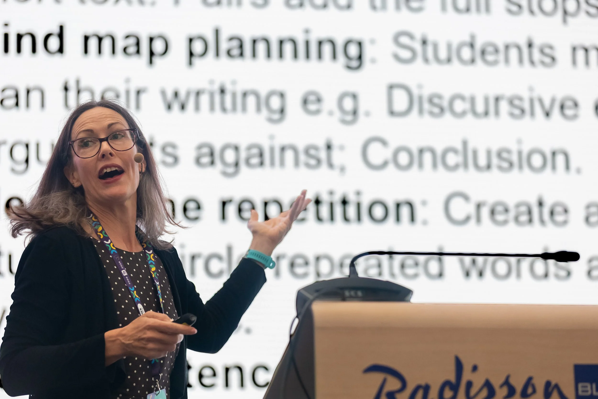 Woman with glasses and long brown hair speaking at a presentation, gesturing with her hand, with a large screen displaying text in the background.