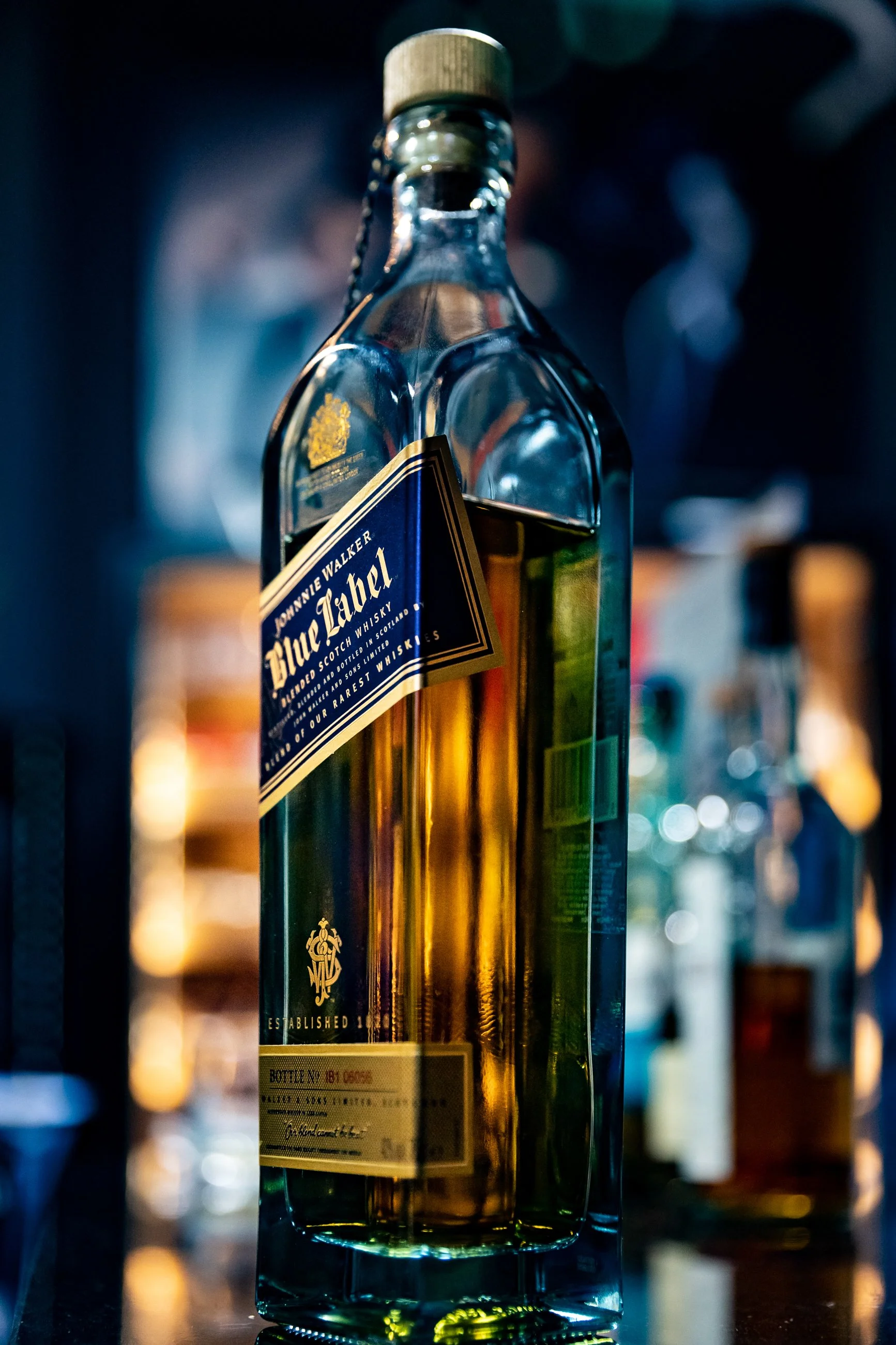 Close-up of a bottle of Johnnie Walker Blue Label Scotch whisky with a striped gold and black label, gold accents, and a wooden cap on a dark reflective surface with bokeh background lights.