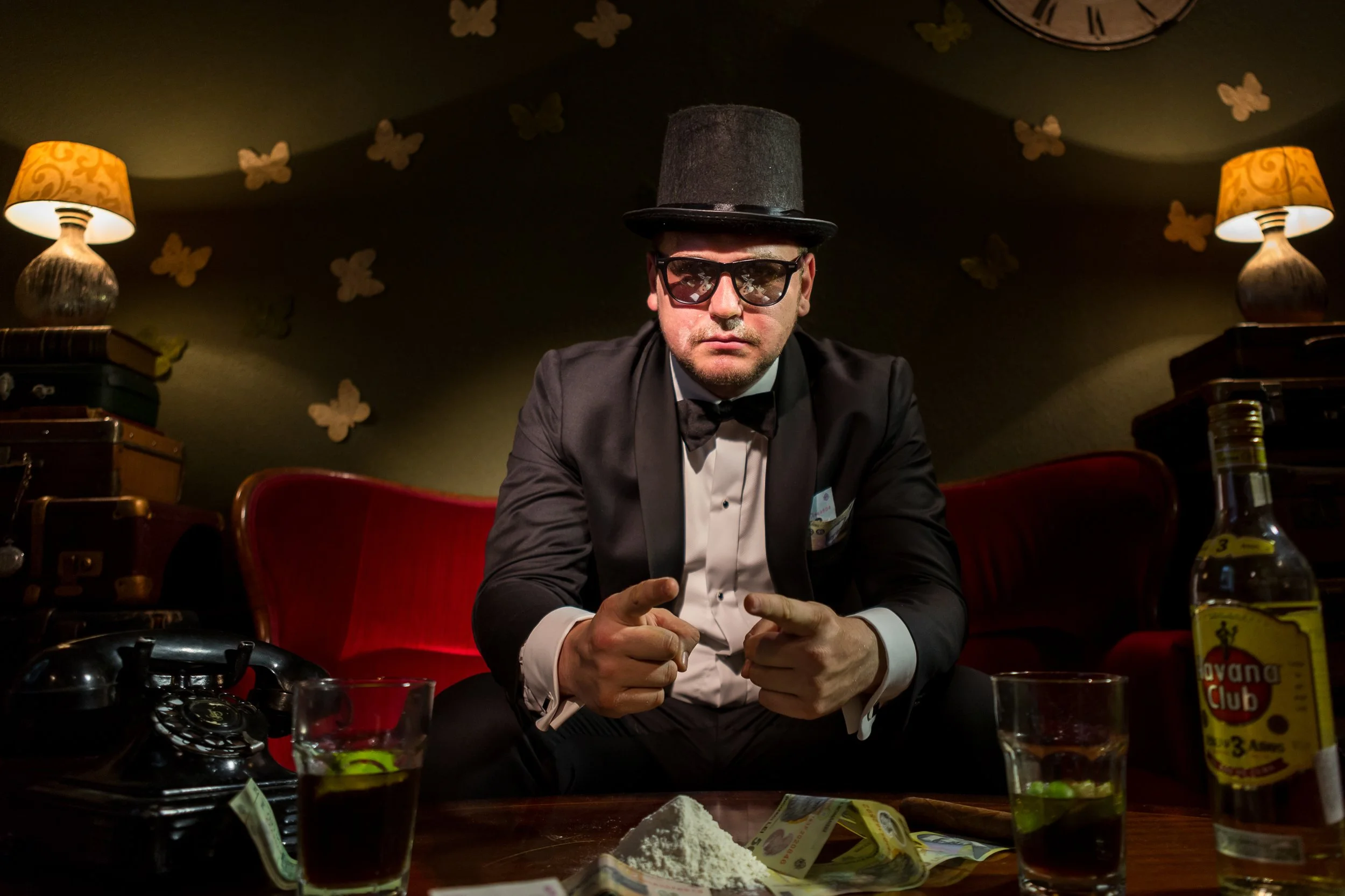 A man dressed in a tuxedo and wearing sunglasses and a tall black hat, sitting at a cluttered table with drinks, snacks, and books, in a room decorated with butterfly wall stickers and lit by two yellow lamps with ornate shades.