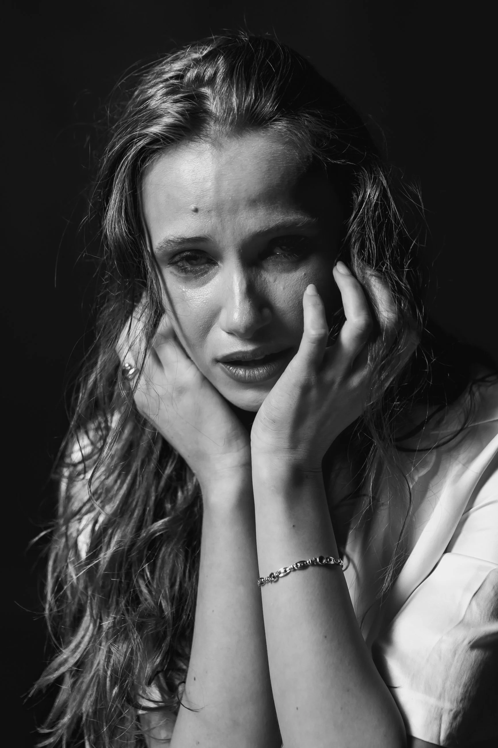 Black and white portrait of a woman with long, wavy hair, looking at the camera with her hand resting on her face, wearing a bracelet and earrings.