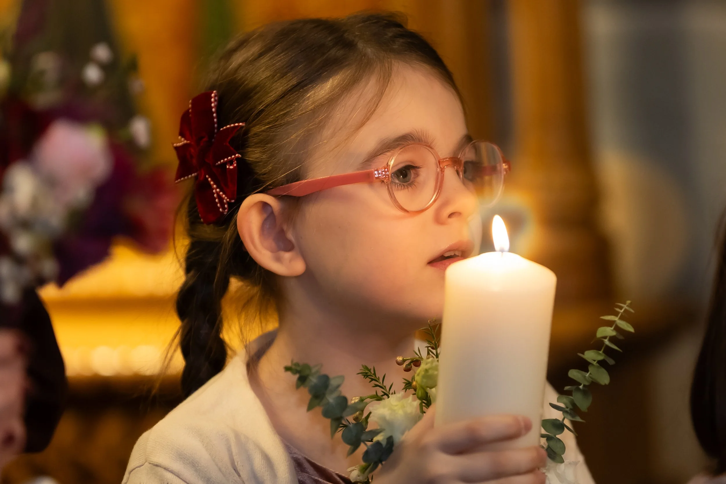 A young girl with glasses and a red bow in her hair holding a lit candle during a holiday celebration.