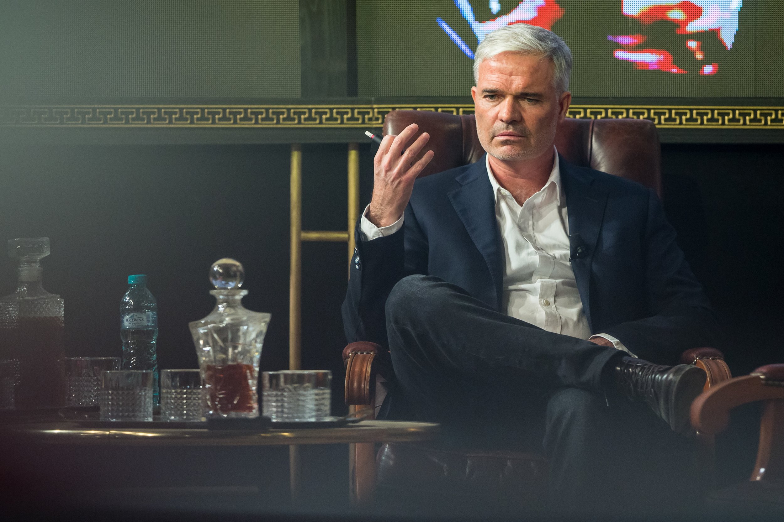 A man with gray hair sitting in a leather armchair, holding a cigarette in his right hand, with a serious expression on his face. Behind him is a screen with a colorful, pixelated map of the world. On the table in front of him are bottles, glasses, a