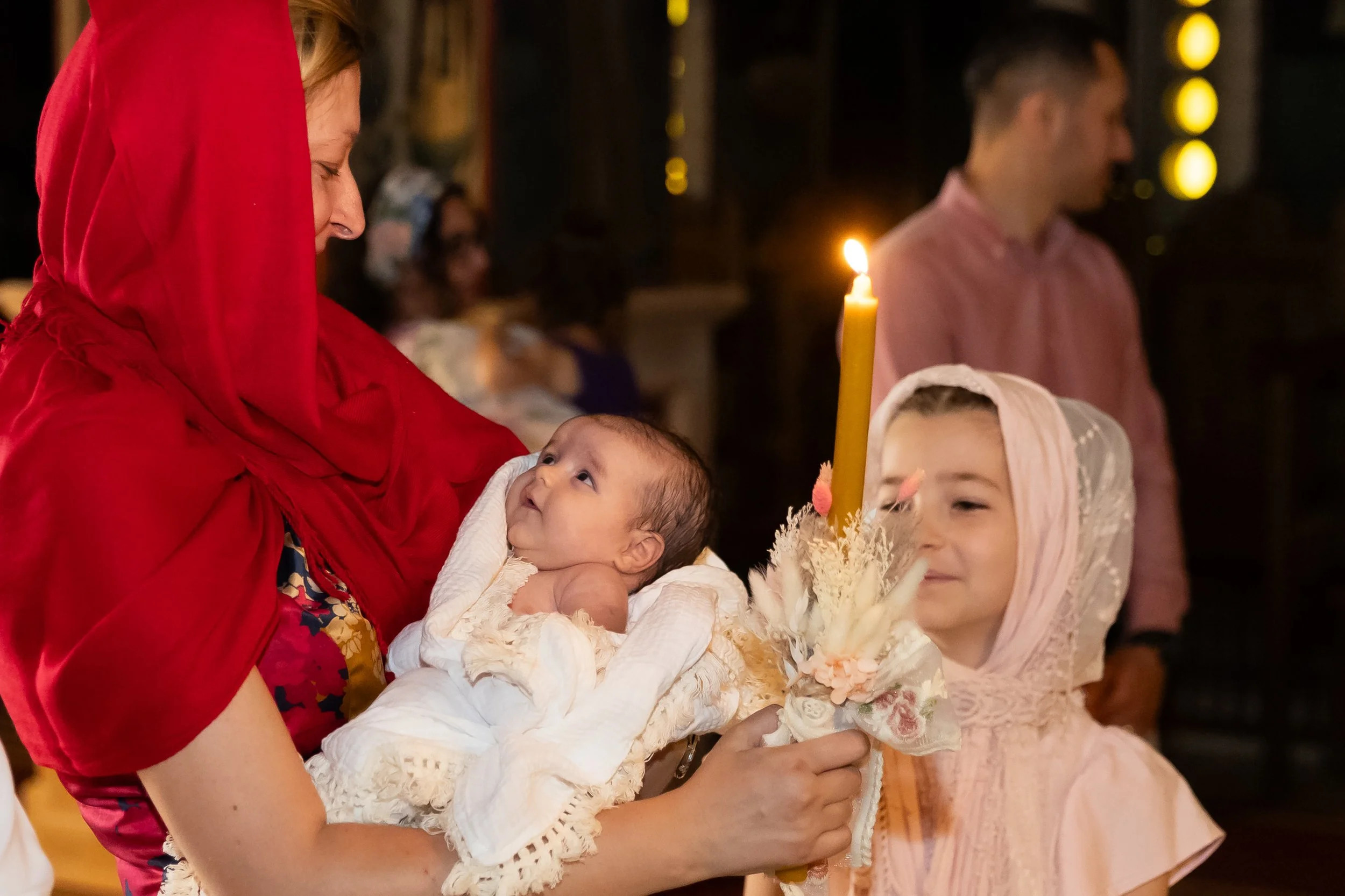 A woman in a red head covering holds a baby dressed in white during a religious ceremony. A girl in pink and an older woman in a pink dress are also participating, holding a candle and flowers in a dimly lit setting.