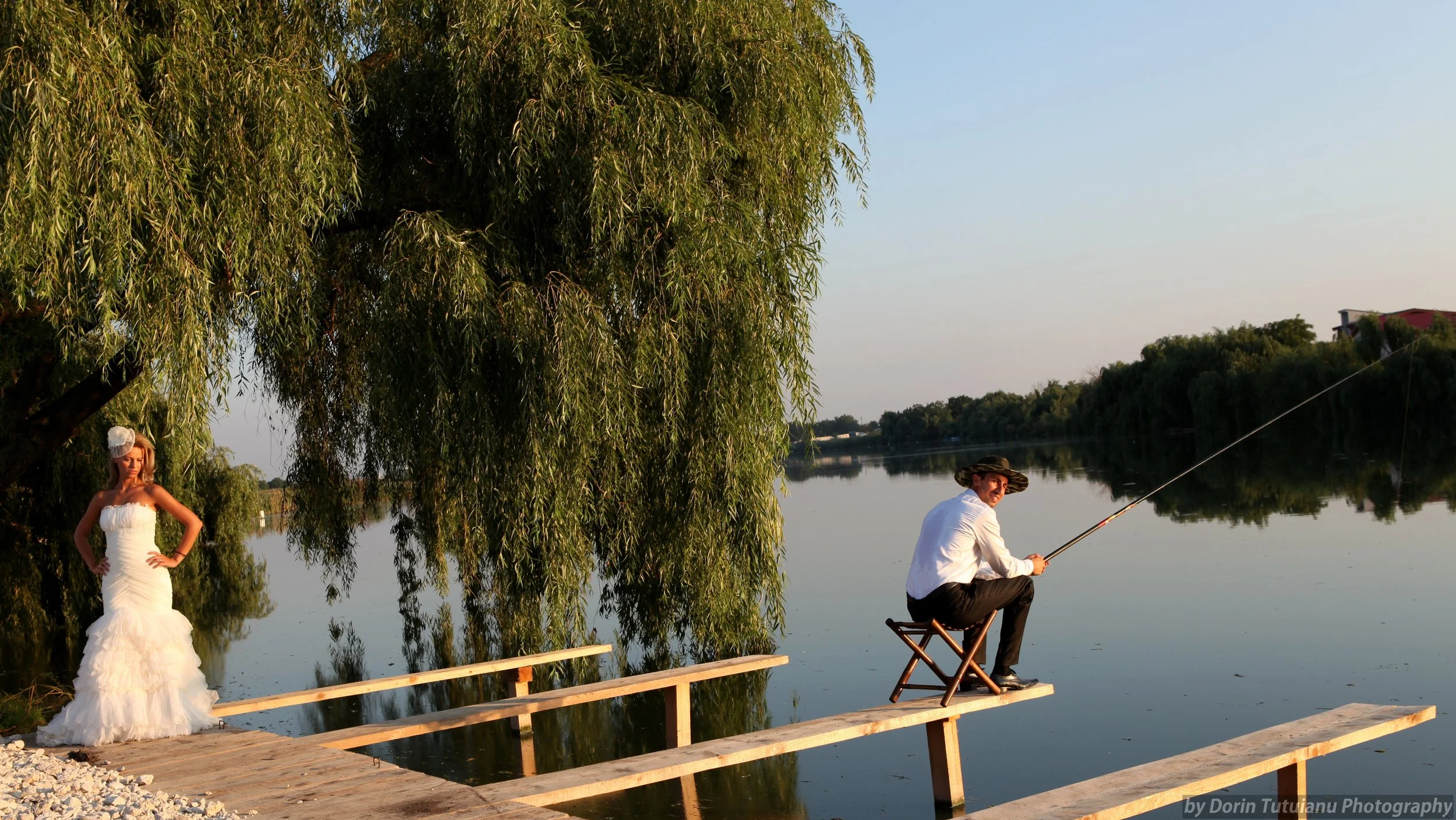 A woman in a white strapless dress posing beside a tree near a river, and a man in a white shirt and hat sitting on a wooden dock fishing by the water during sunset.