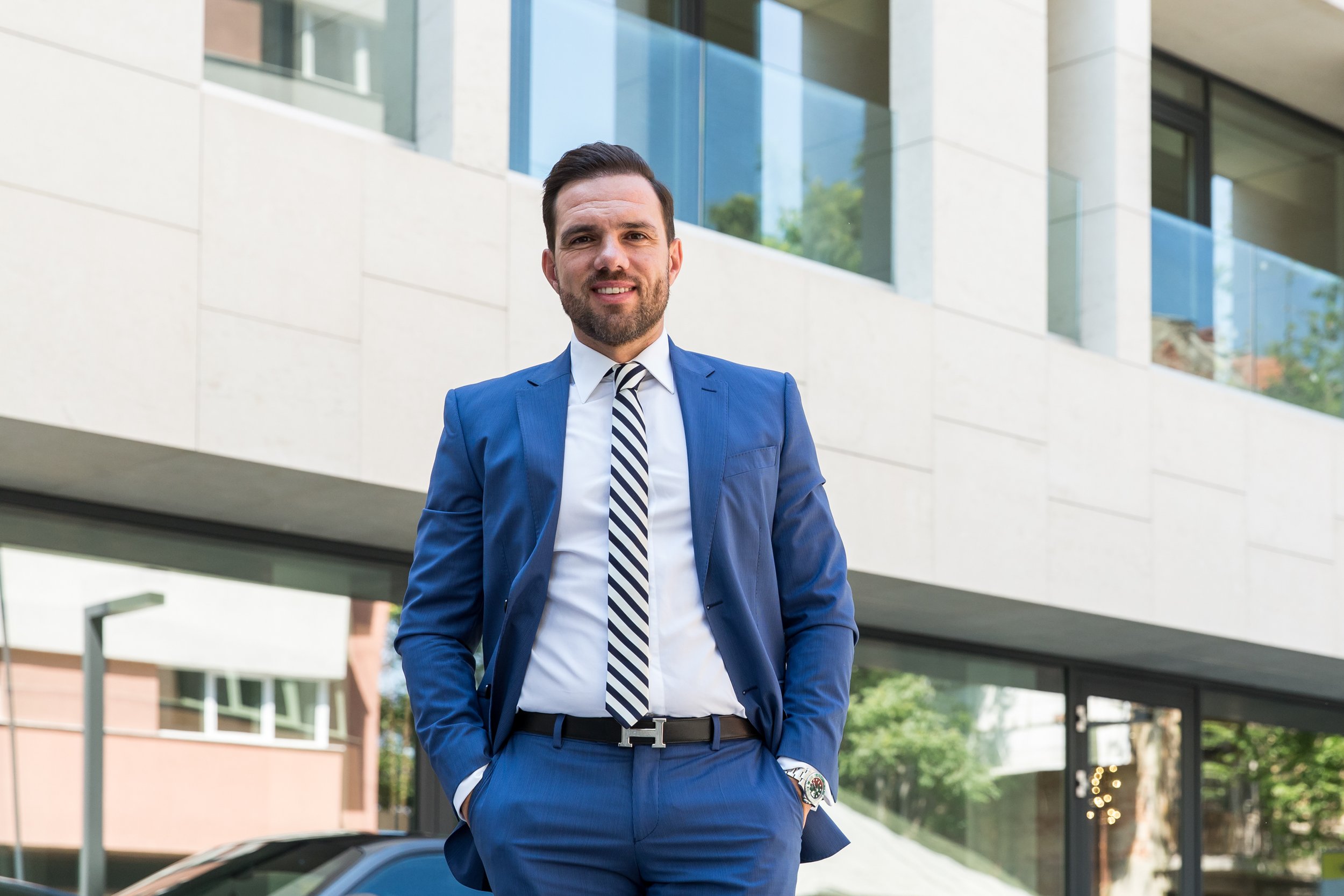 A man wearing a blue suit, striped tie, and white shirt standing outdoors in front of a modern building, smiling at the camera.