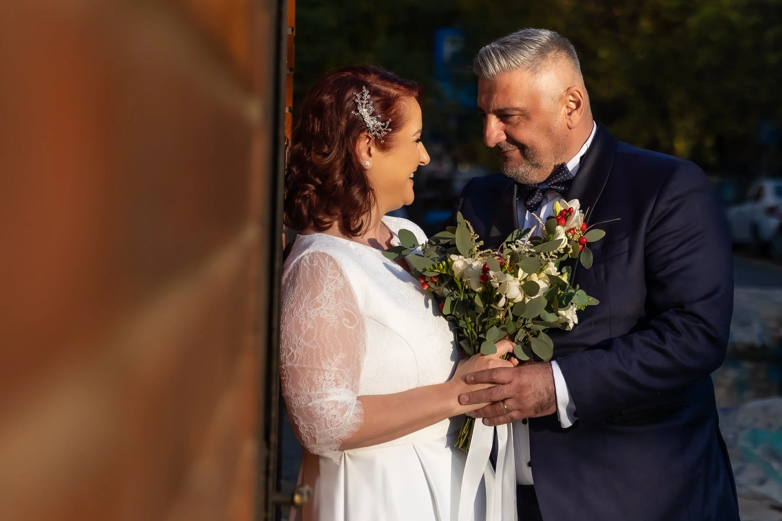 A bride and groom facing each other with foreheads touching, holding hands, outdoors during sunset, the bride with a bouquet and the groom in a tuxedo with a bow tie.