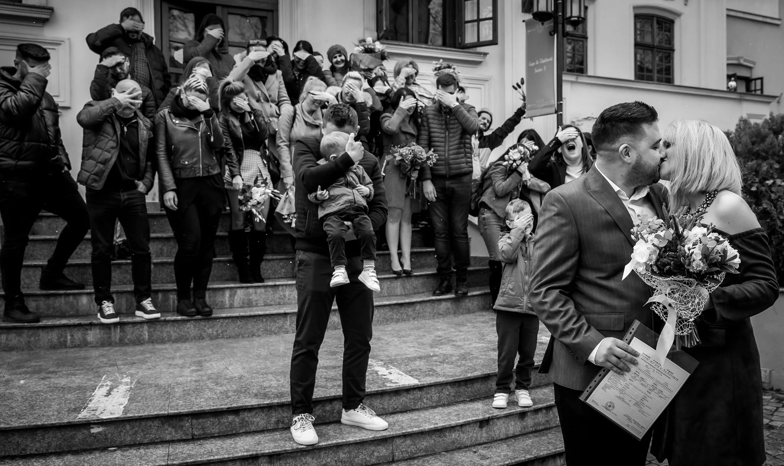 A group of people on steps, covering their eyes, with two individuals in the foreground kissing, one holding a bouquet and a certificate, in black and white.
