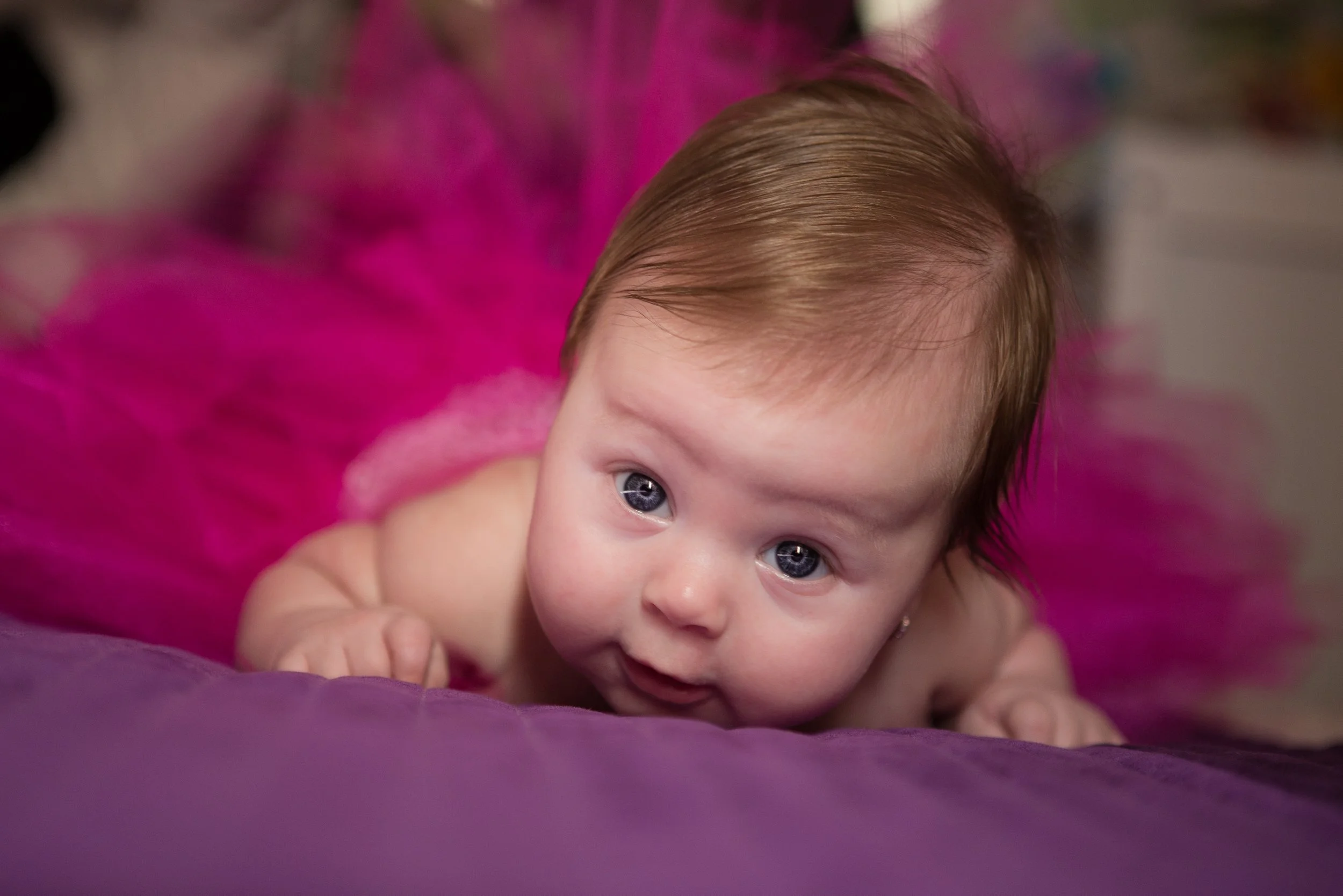 Close-up of a baby with blue eyes and light brown hair, lying on a purple surface with pink feather-like decoration in the background.