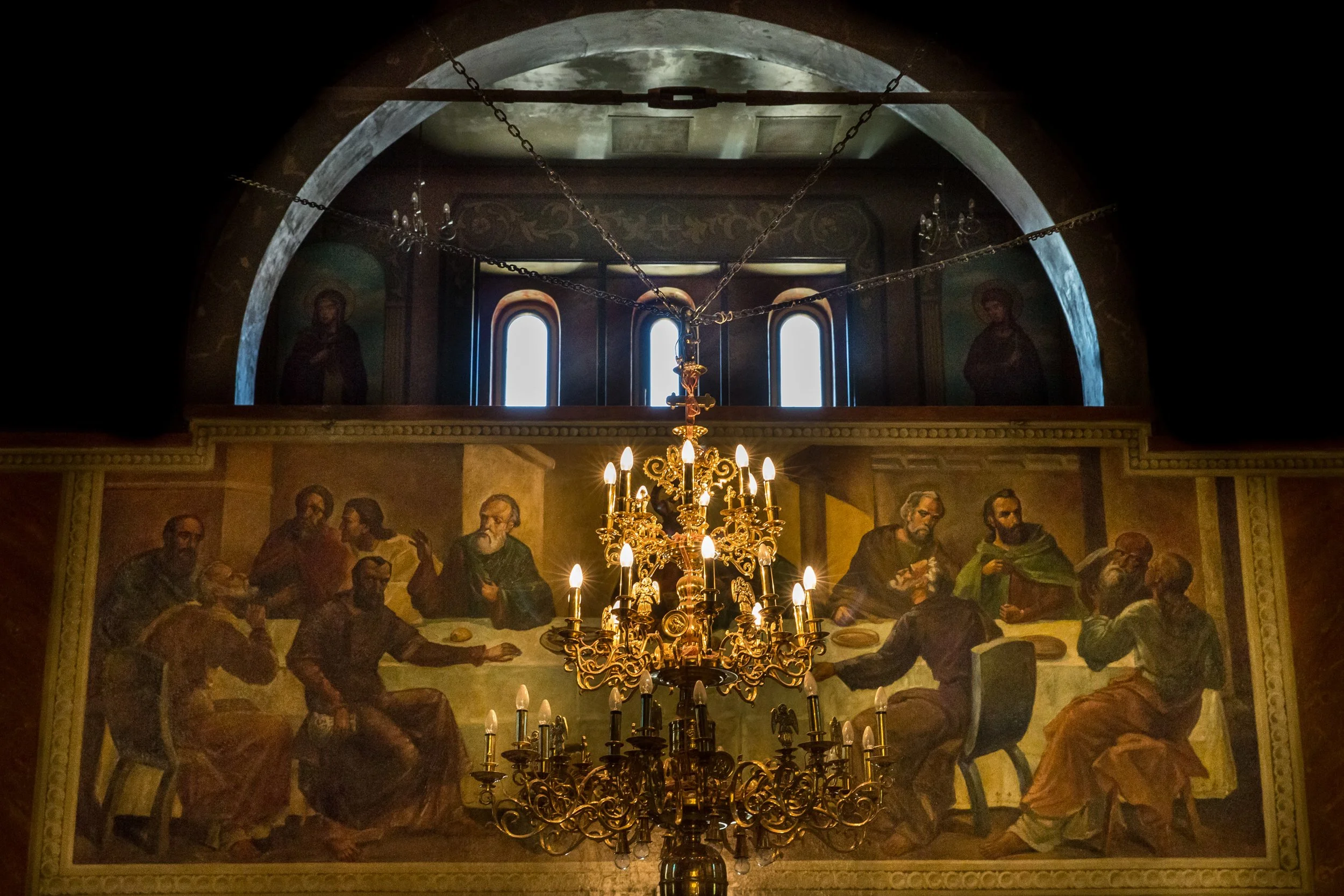 Interior of a church or cathedral with a chandelier in the foreground, a religious mural or painting depicting biblical figures, and windows above the mural.