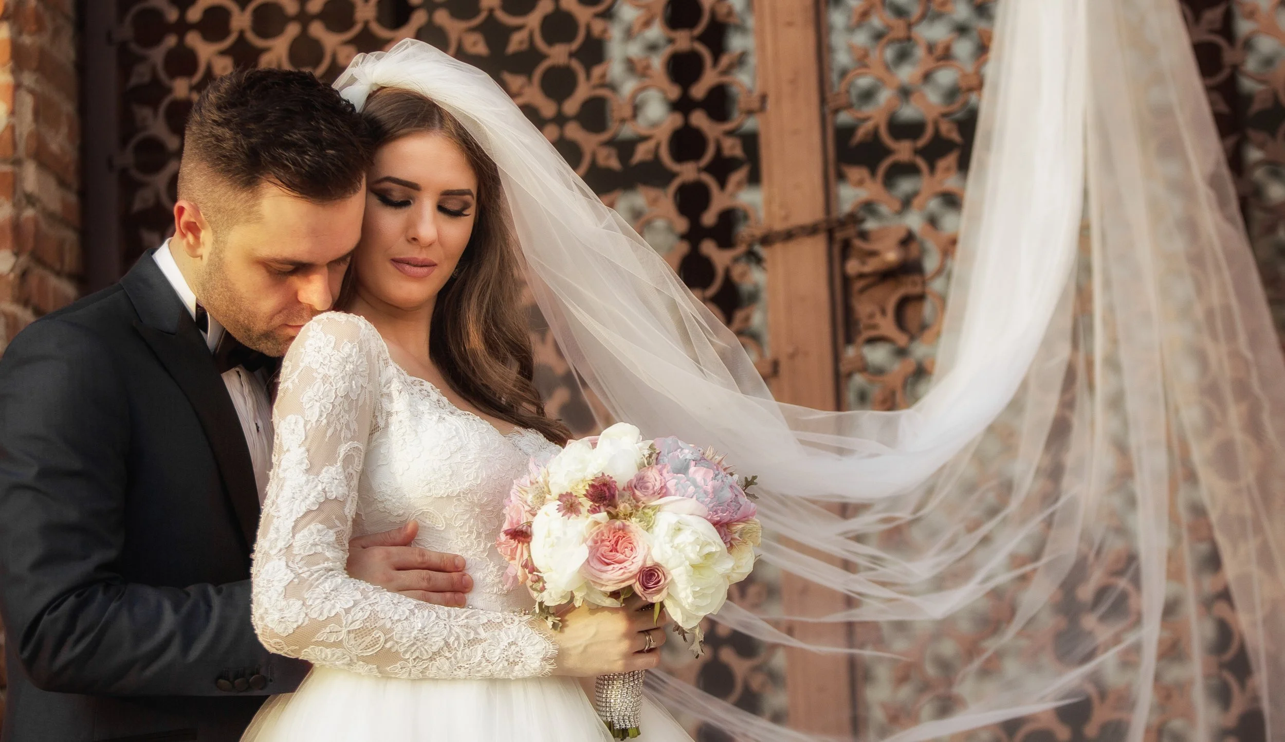 A bride and groom share an intimate moment, with the groom's head resting against the bride's forehead. The bride is holding a bouquet of pink and white flowers, and both are dressed in formal wedding attire. The background features a brick wall and 