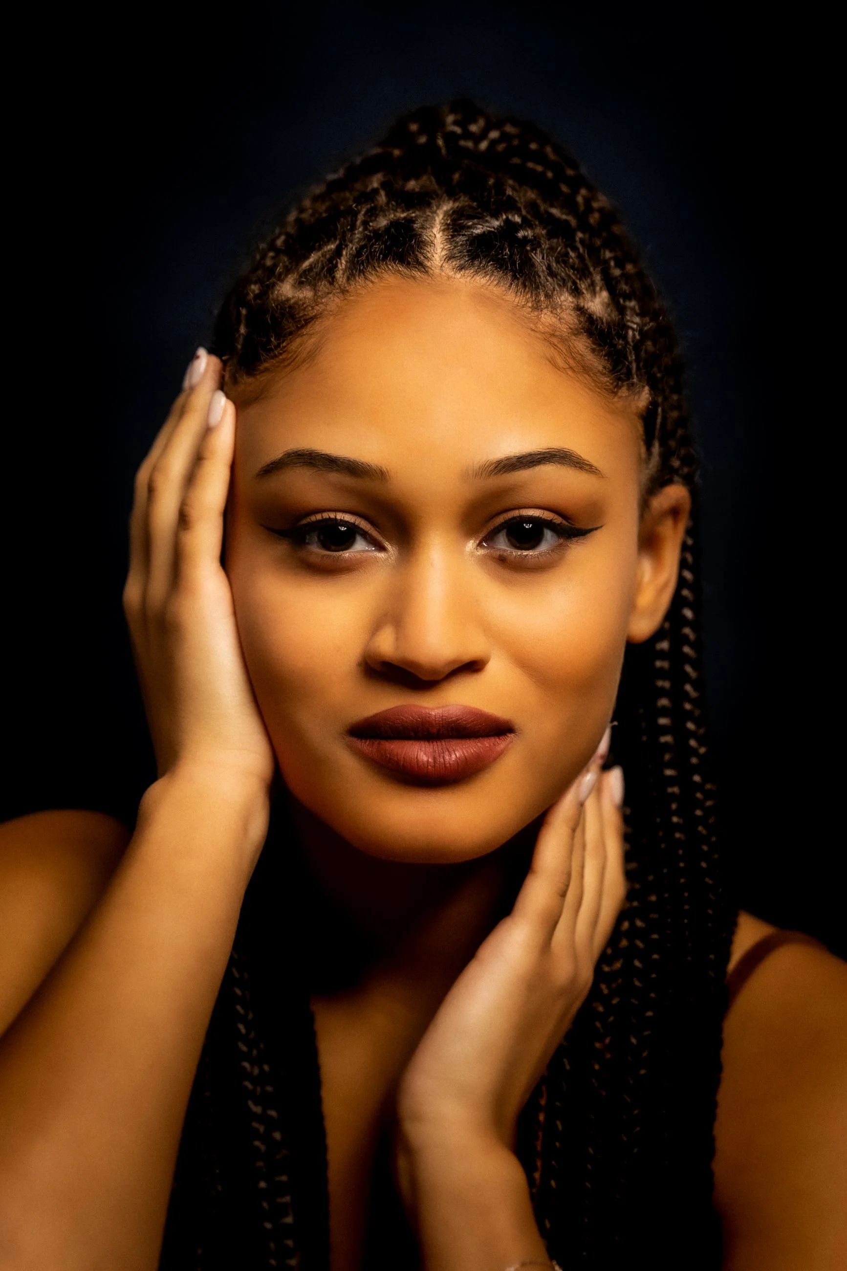 Close-up portrait of a young woman with braided hair, possibly in a studio setting with dark background, touching her face gently with both hands.