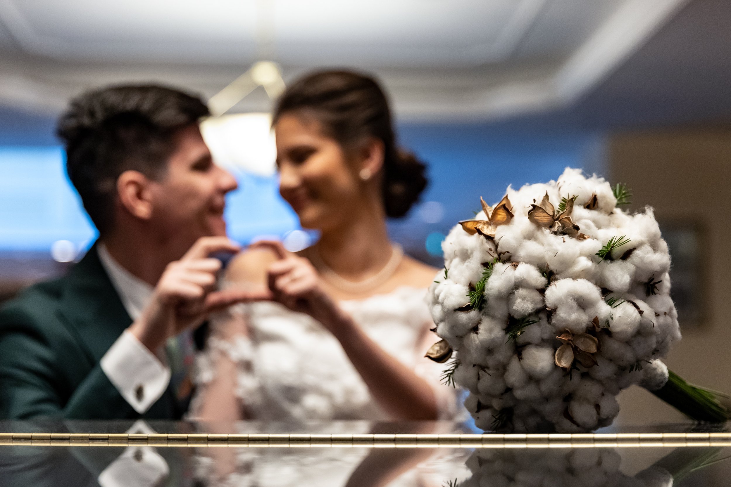 A man and woman, dressed in formal wedding attire, are taking a photo or video together, smiling and leaning close. In the foreground, a large bouquet of cotton flowers with brown Butterfly butterflies is visible.