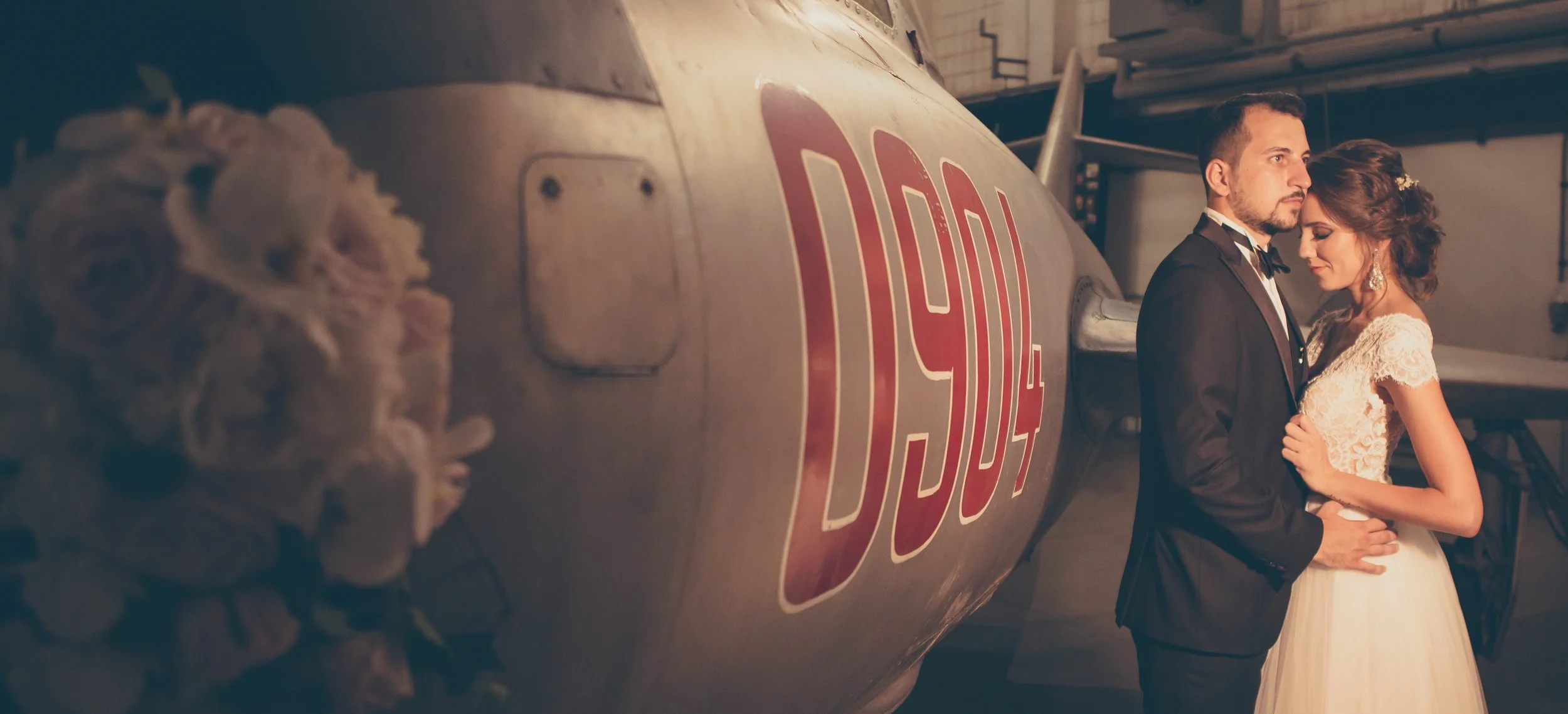 A bride and groom standing close together in front of an airplane with the markings 'DC-3' and a registration number. The bride is wearing a lace wedding dress, and the groom is in a tuxedo. They are indoors, with the airplane in the background and s