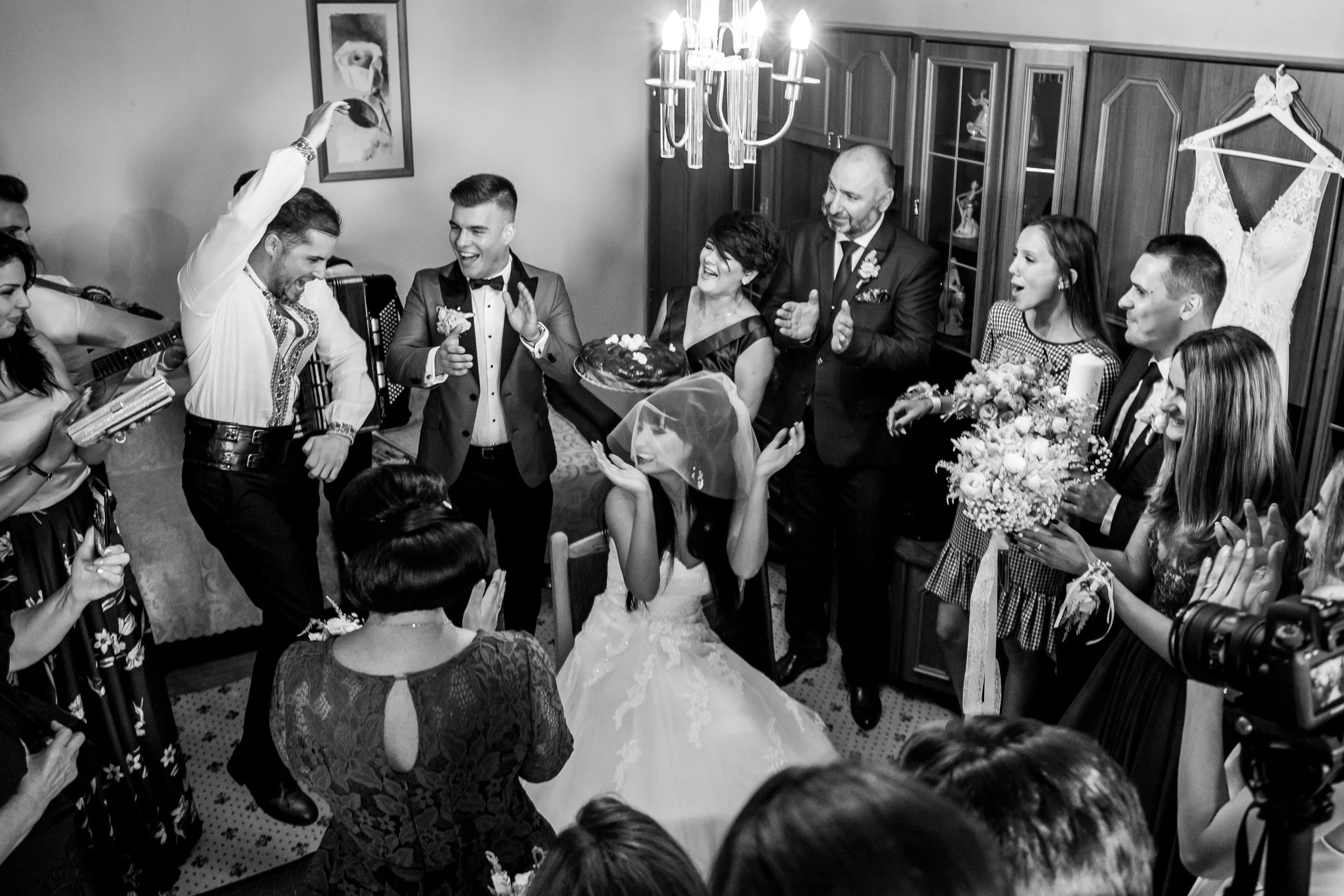 A black and white photo of a wedding celebration with a bride and groom at the center, surrounded by smiling guests. The groom is holding a cake, and the bride is sitting on a chair. Guests are clapping and celebrating, with one person pouring a drin