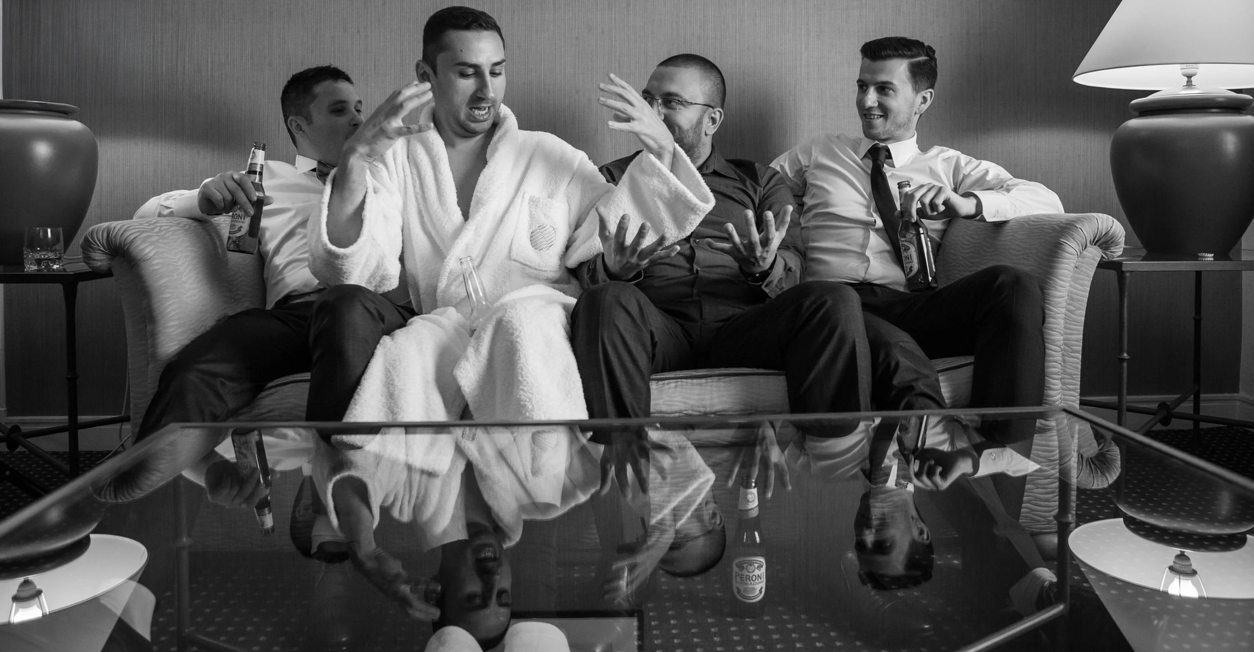 Four men sitting on a sofa, with three holding bottles of beer, one wearing a bathrobe, engaged in conversation in a living room with lamps and a glass table, in black and white.